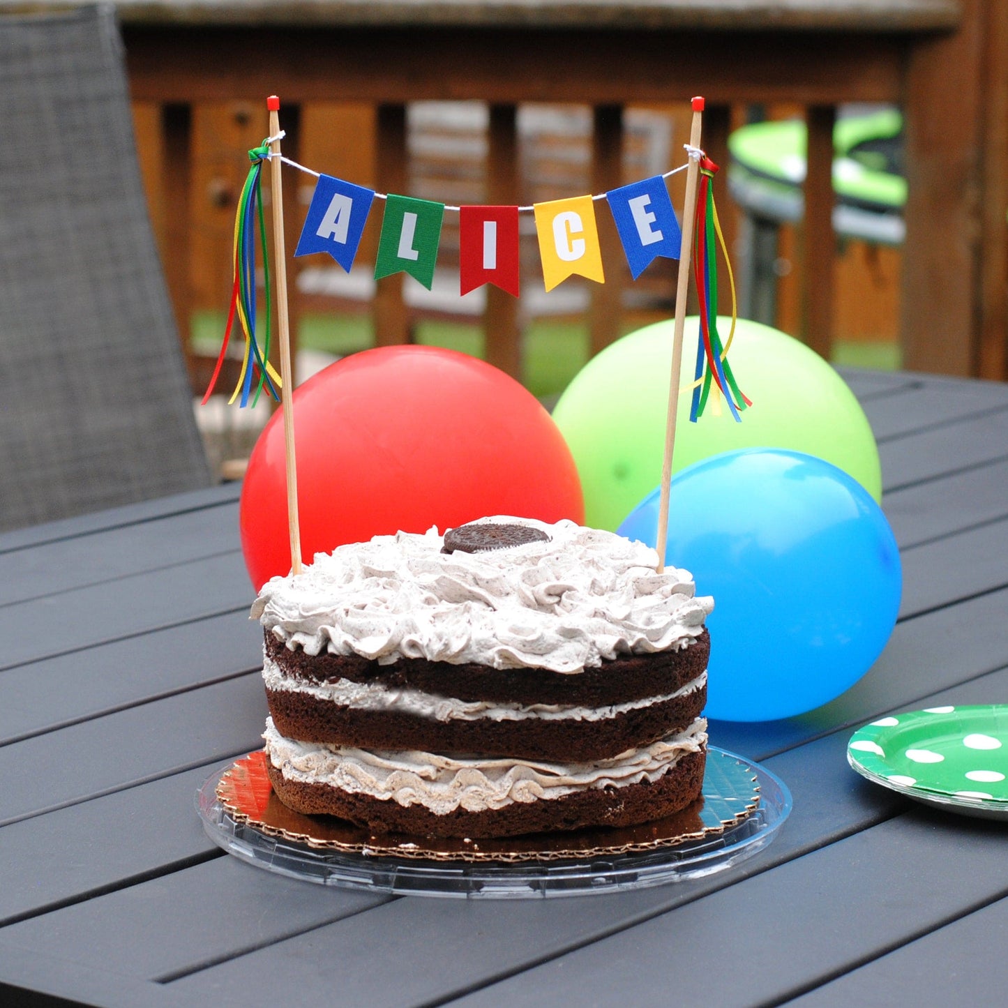 Chocolate cake with white frosting on a table outdoors, decorated with colorful balloons and a 'ALICE' personalized cake topper in bright colors | personalized cake toppers by Avalon Sunshine