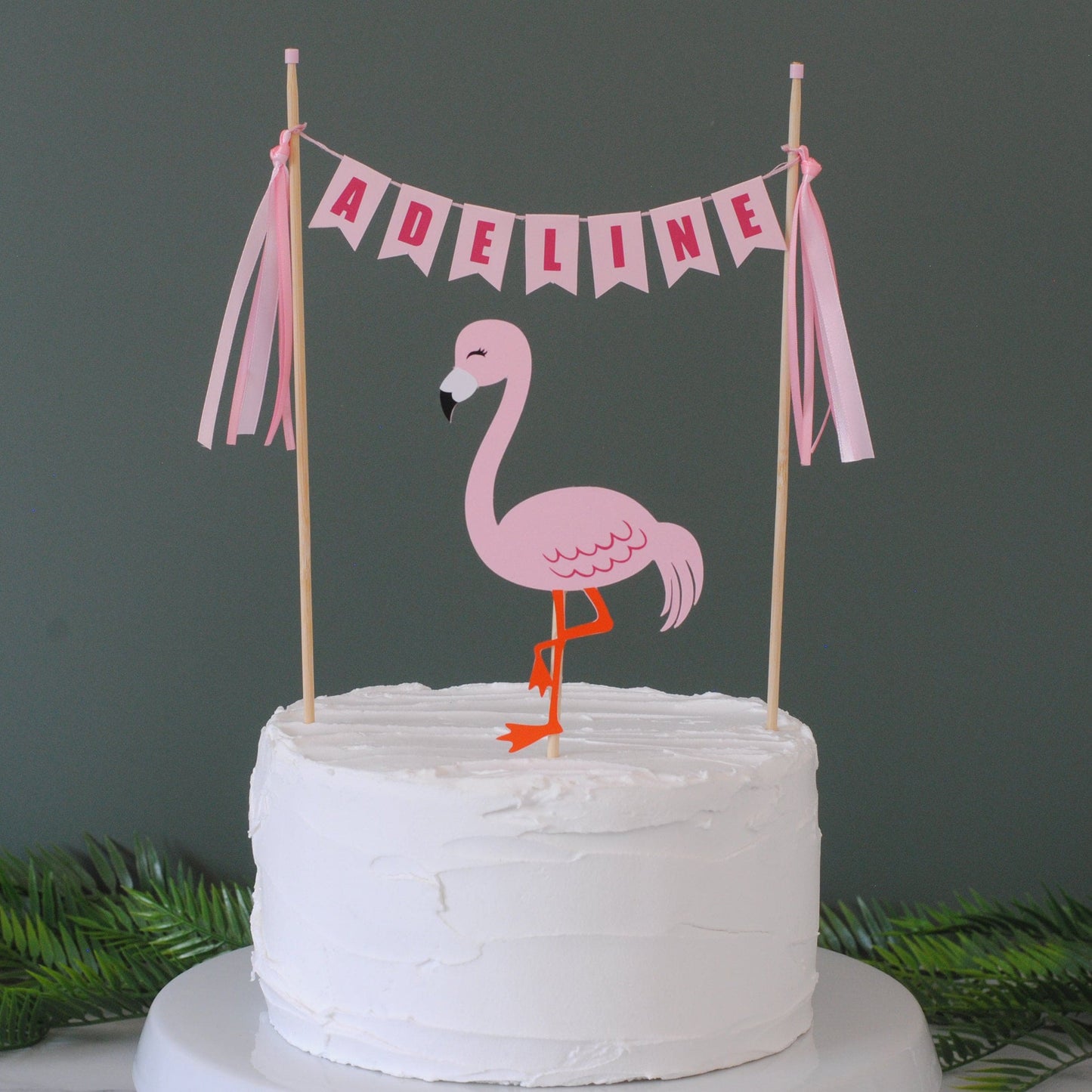 Pink flamingo cake topper set including a pink personalized name banner with pink ribbon tassels.  Shown on a white cake with tropical greenery in the background | personalized cake toppers by Avalon Sunshine