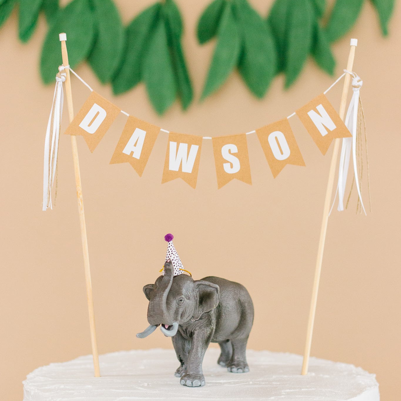 neutral light brown personalized cake topper with name shown on child's birthday cake with a toy elephant