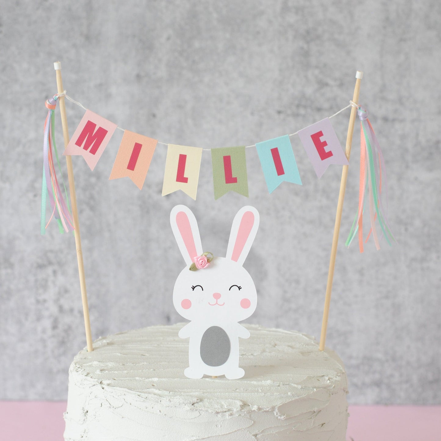 pastel rainbow name banner personalized with name.  Paper bunny cake topper underneath with a flower on its ear.