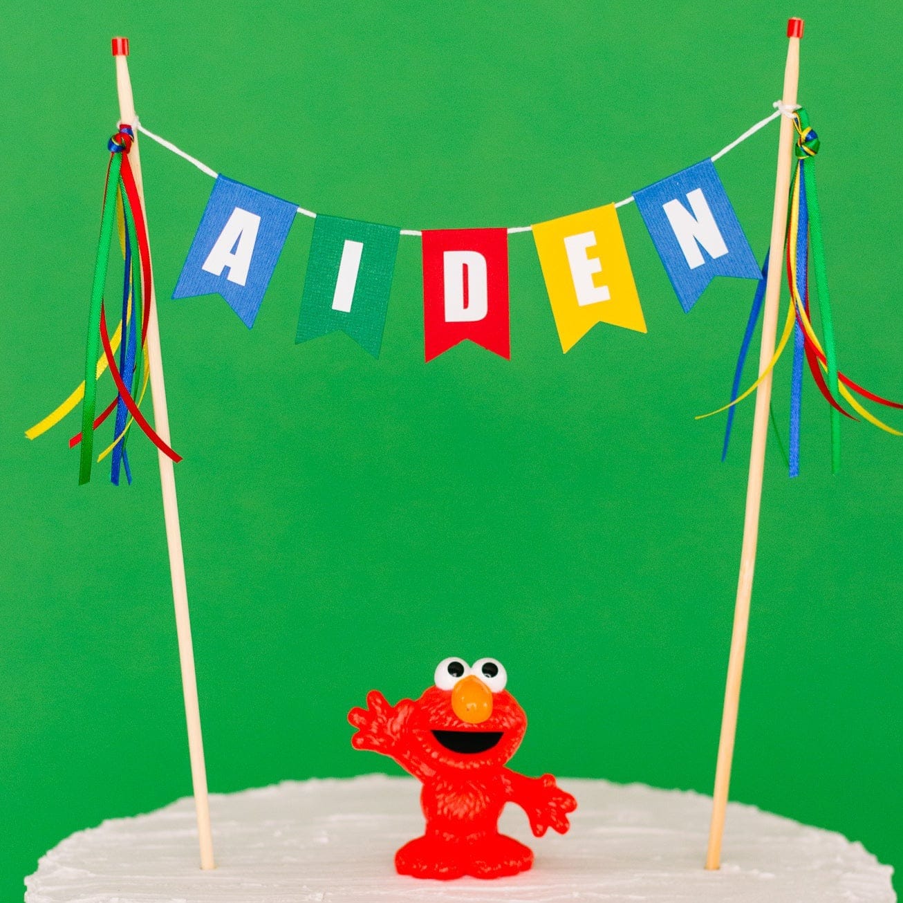 
                  
                    multi-color personalized name cake topper in blue, green, red and yellow shown with an Elmo figure on top of the cake
                  
                