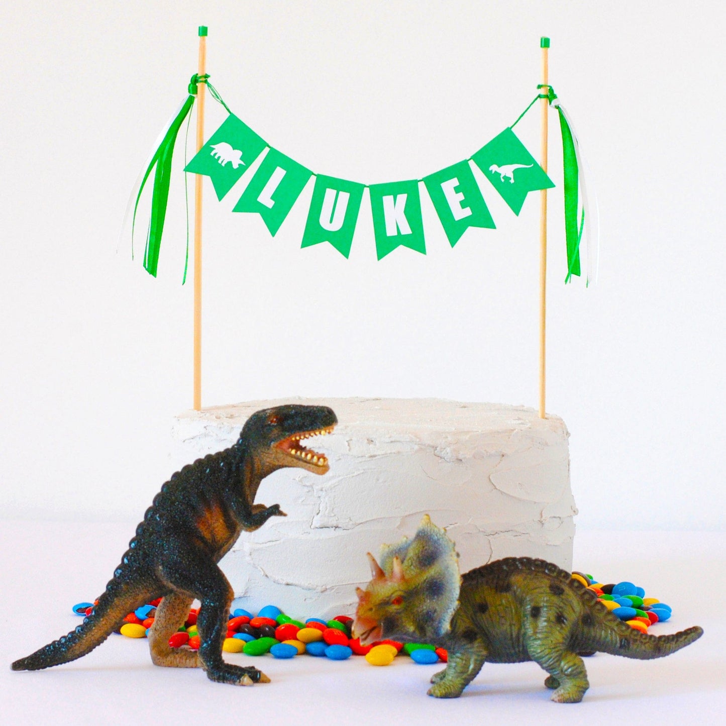 White birthday cake with green dinosaur theme cake banner and toy dinosaur decorations