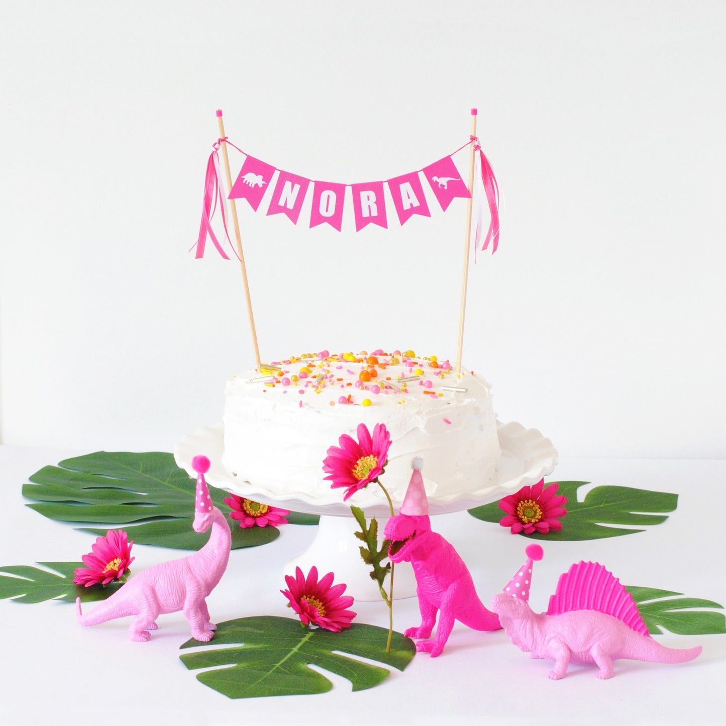 white birthday cake with bright dinosaur theme name banner on top.  Pink dinosaur decorations and tropical leaves as table decorations