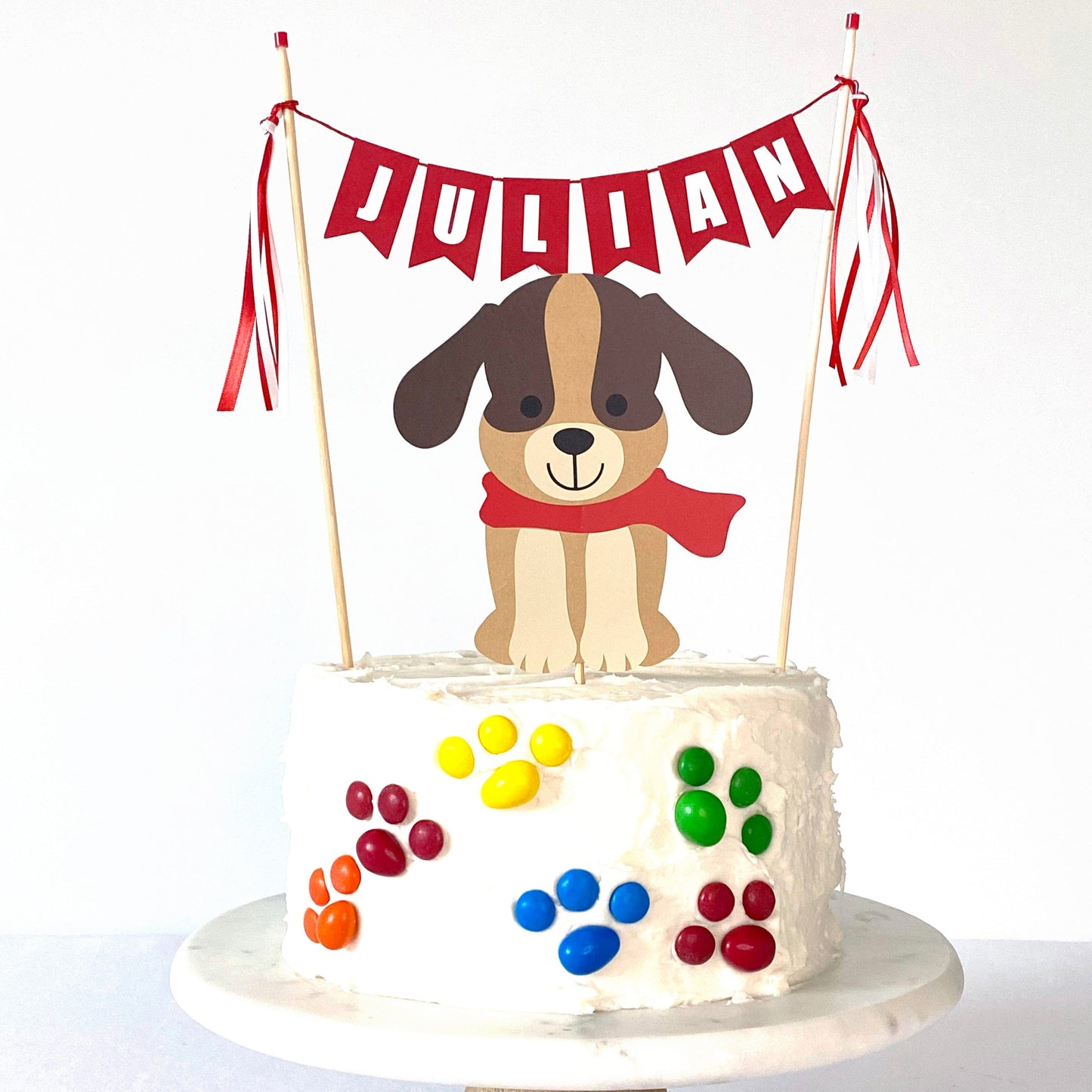 
                  
                    dog theme birthday cake topper set with brown dog wearing a red scarf and red name cake banner on a white cake decorated with M&M candies in paw print shapes
                  
                