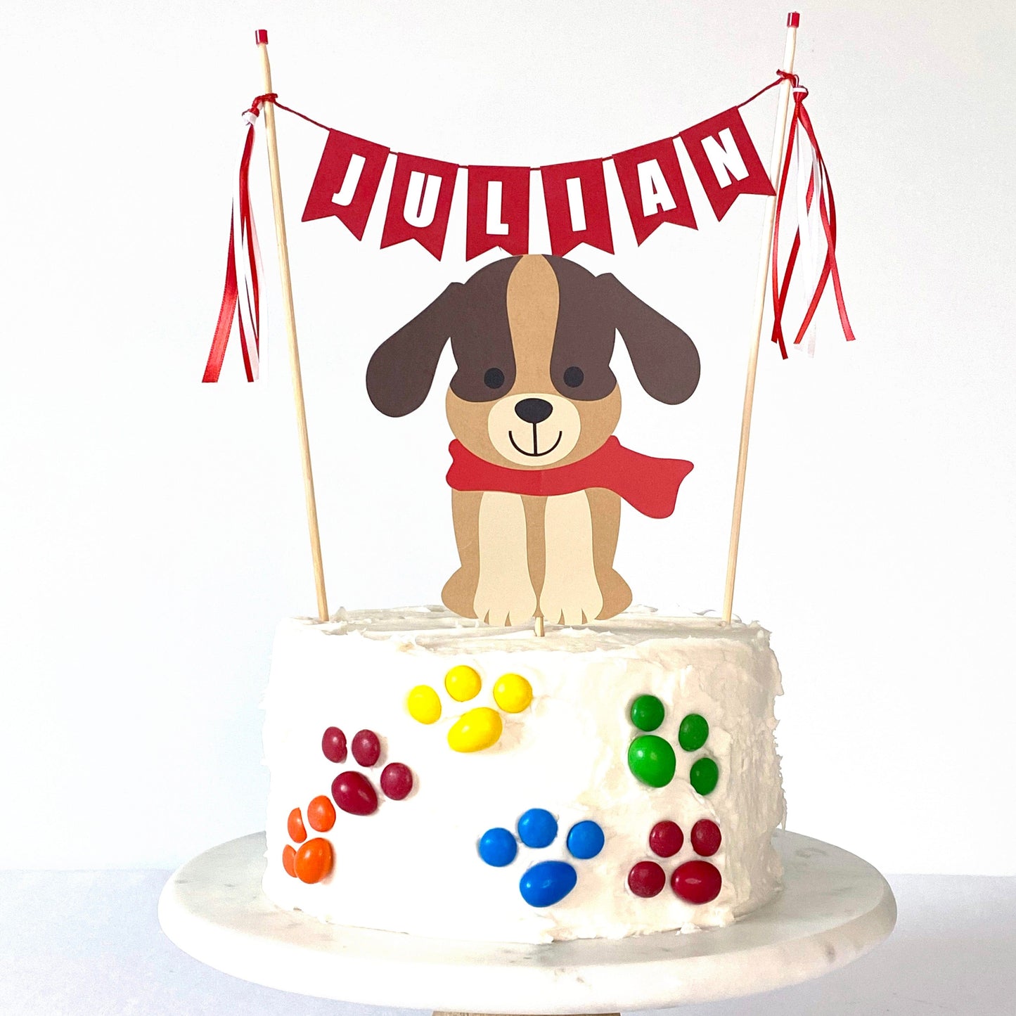 dog theme birthday cake topper set with brown dog wearing a red scarf and red name cake banner on a white cake decorated with M&M candies in paw print shapes
