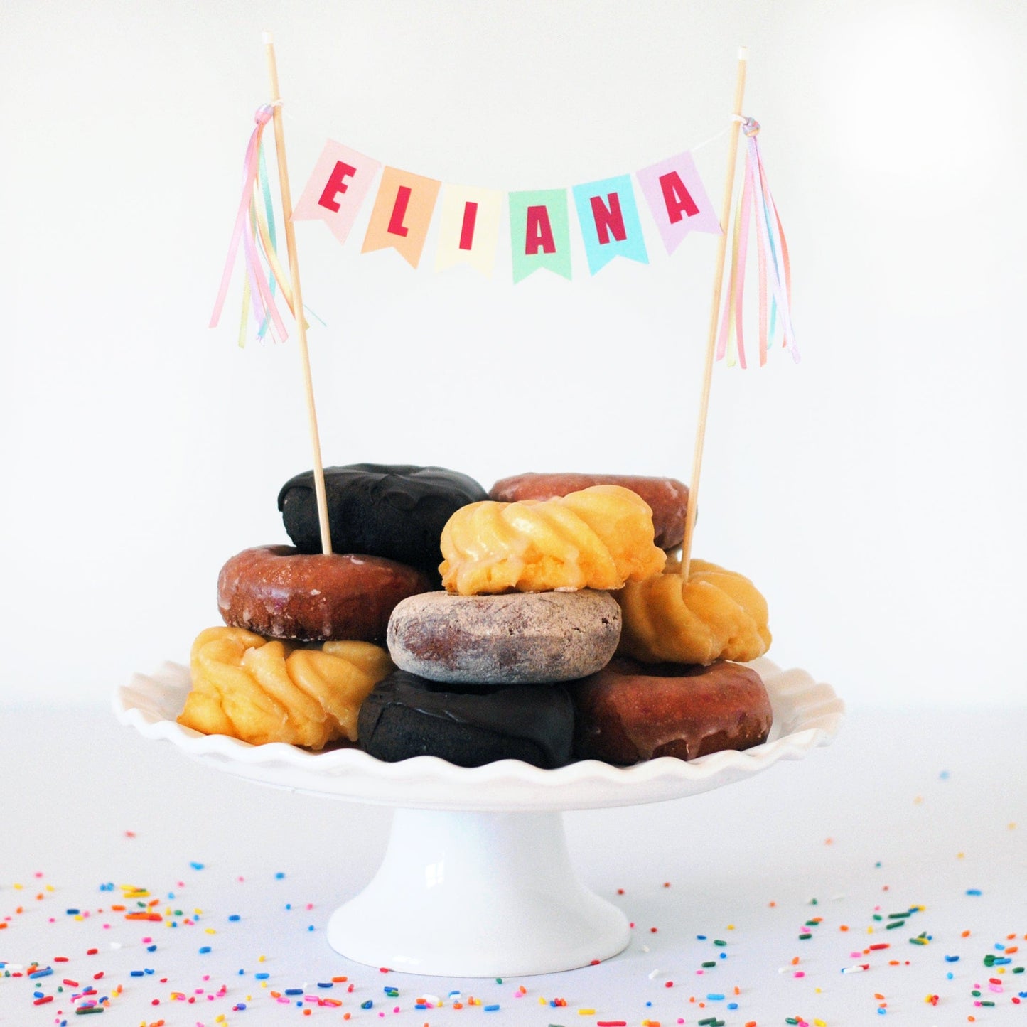 
                  
                    pastel rainbow name cake topper shown on donuts stacked on a cake stand
                  
                