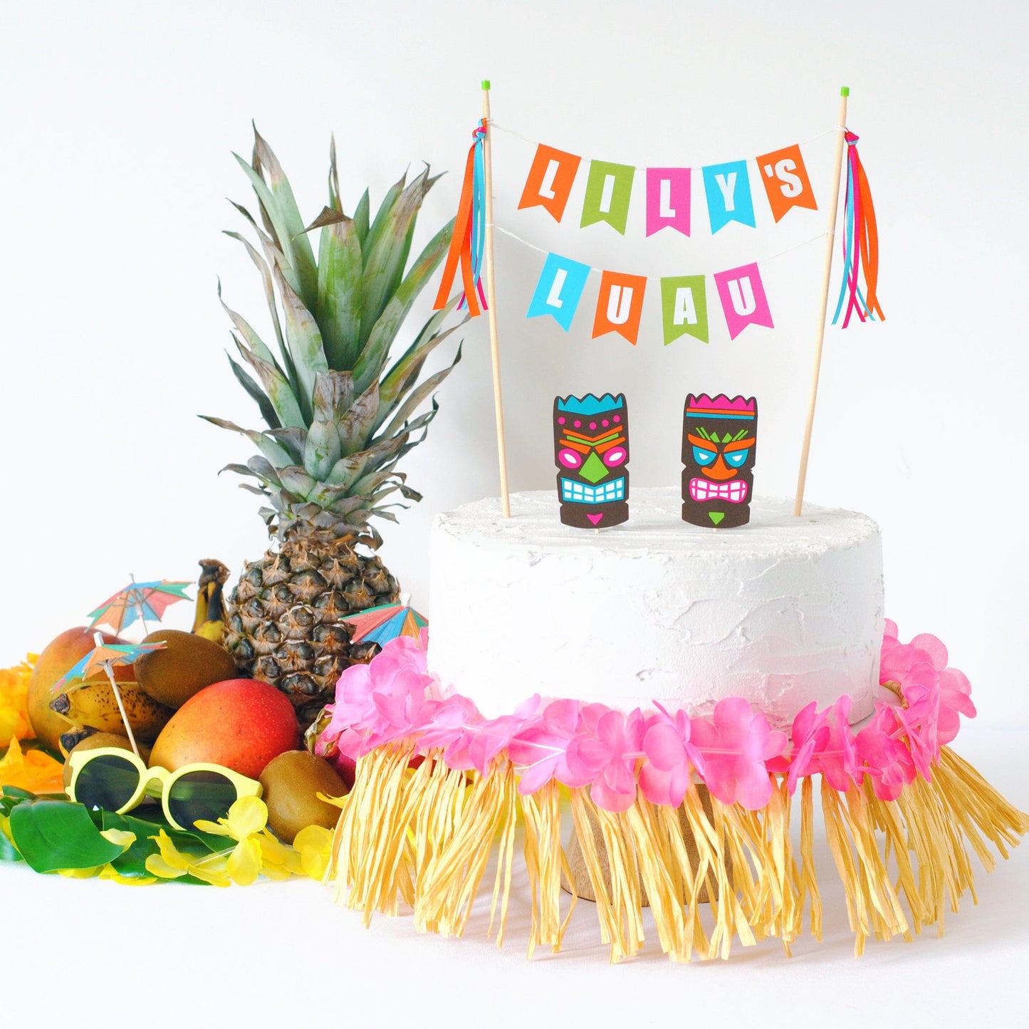 Hawaiian cake topper personalized with name and tiki masks. Table decorations include grass skirt and tropical fruit