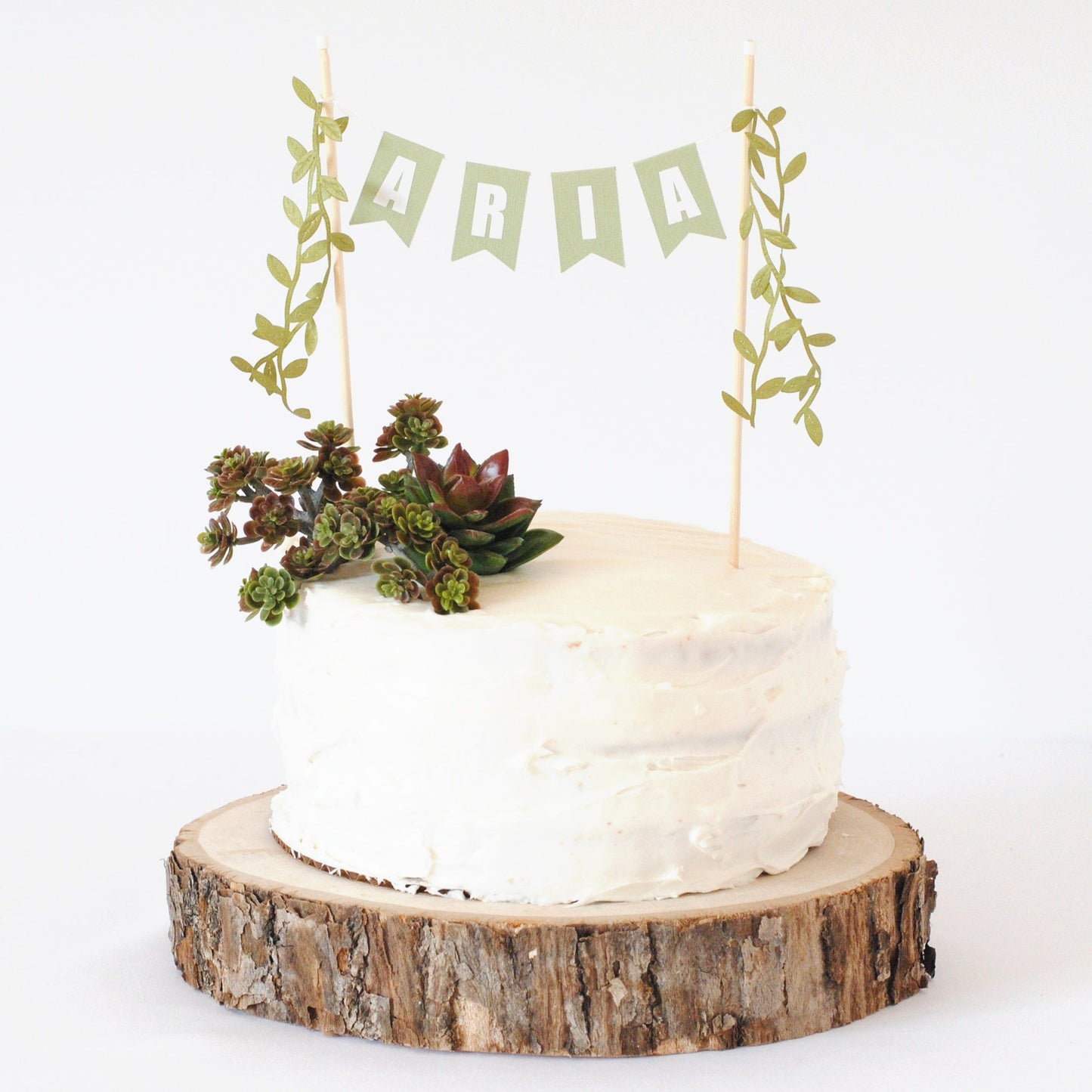 Name cake topper with green leaf ribbons on the ends shown on a white cake with succulents