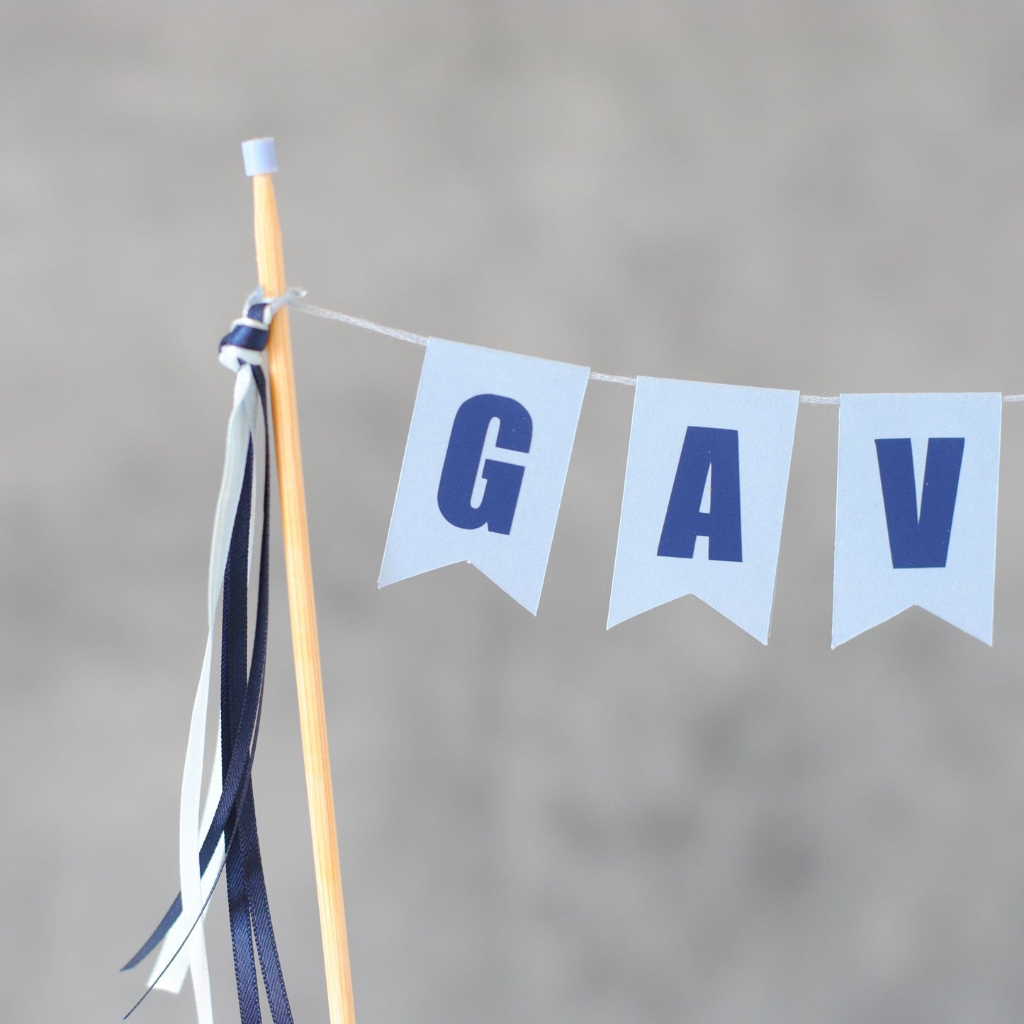 close-up of personalized cake banner with name, dark blue letters on a light blue background and matching ribbon tassels