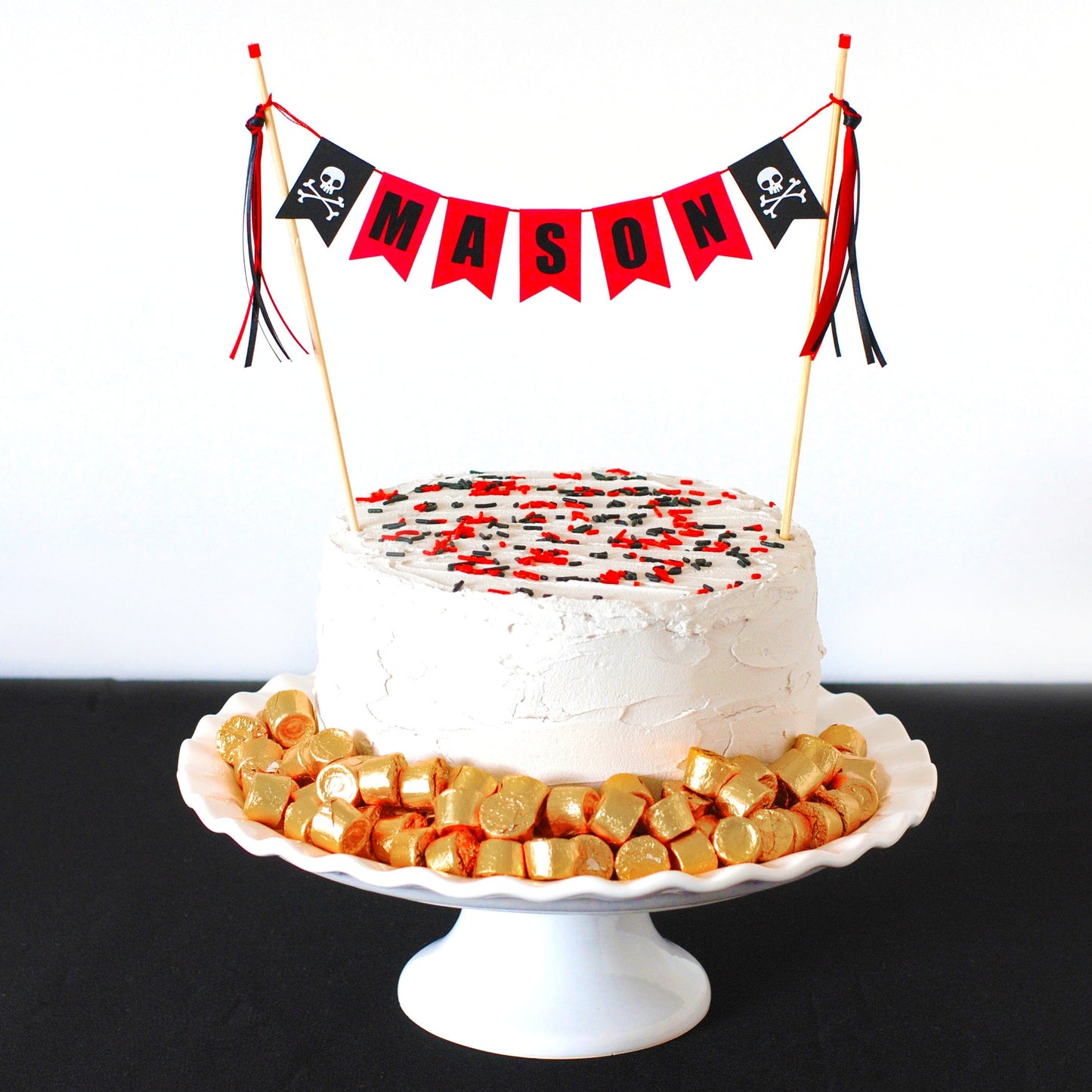 pirate birthday cake topper with name banner and skull and crossbones on each end shown on a white cake with sprinkles and gold rolos