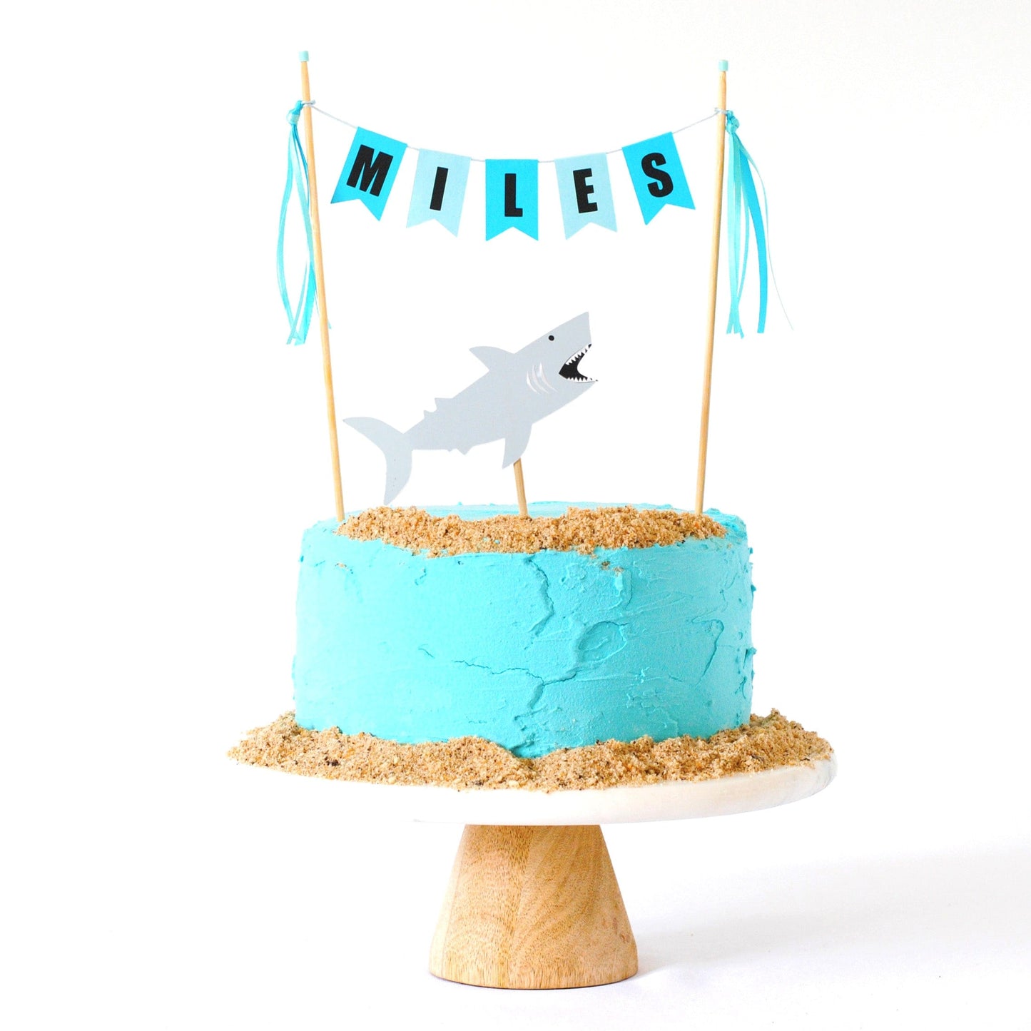 shark birthday cake topper with name banner and shark topper shown on a blue cake with crumbled cookies to look like sand around the cake