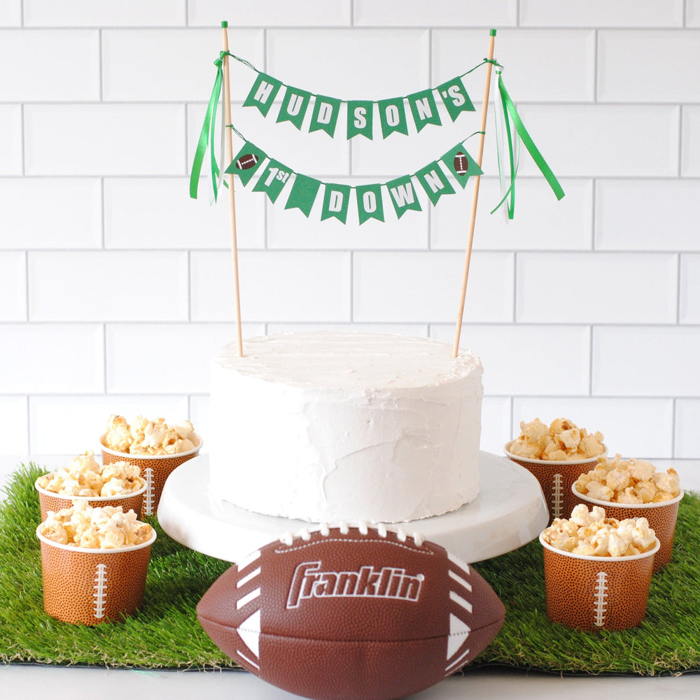 Decorative setup with a cake, popcorn, and football on a grass-like surface against a white tiled wall. Cake has a personalized cake topper saying HUDSON'S 1ST DOWN with footballs on each end | personalized cake toppers by Avalon Sunshine
