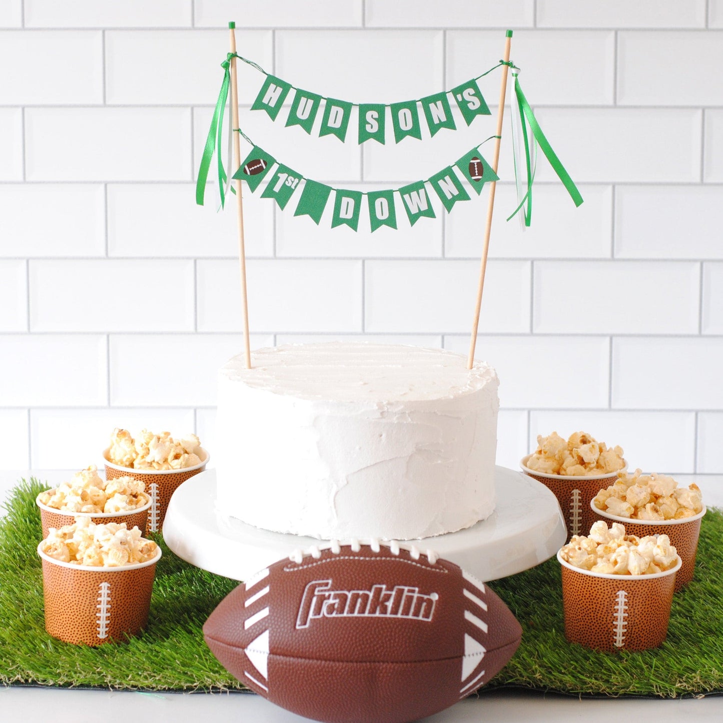 Decorative setup with a cake, popcorn, and football on a grass-like surface against a white tiled wall. Cake has a personalized cake topper saying HUDSON'S 1ST DOWN with footballs on each end | personalized cake toppers by Avalon Sunshine
