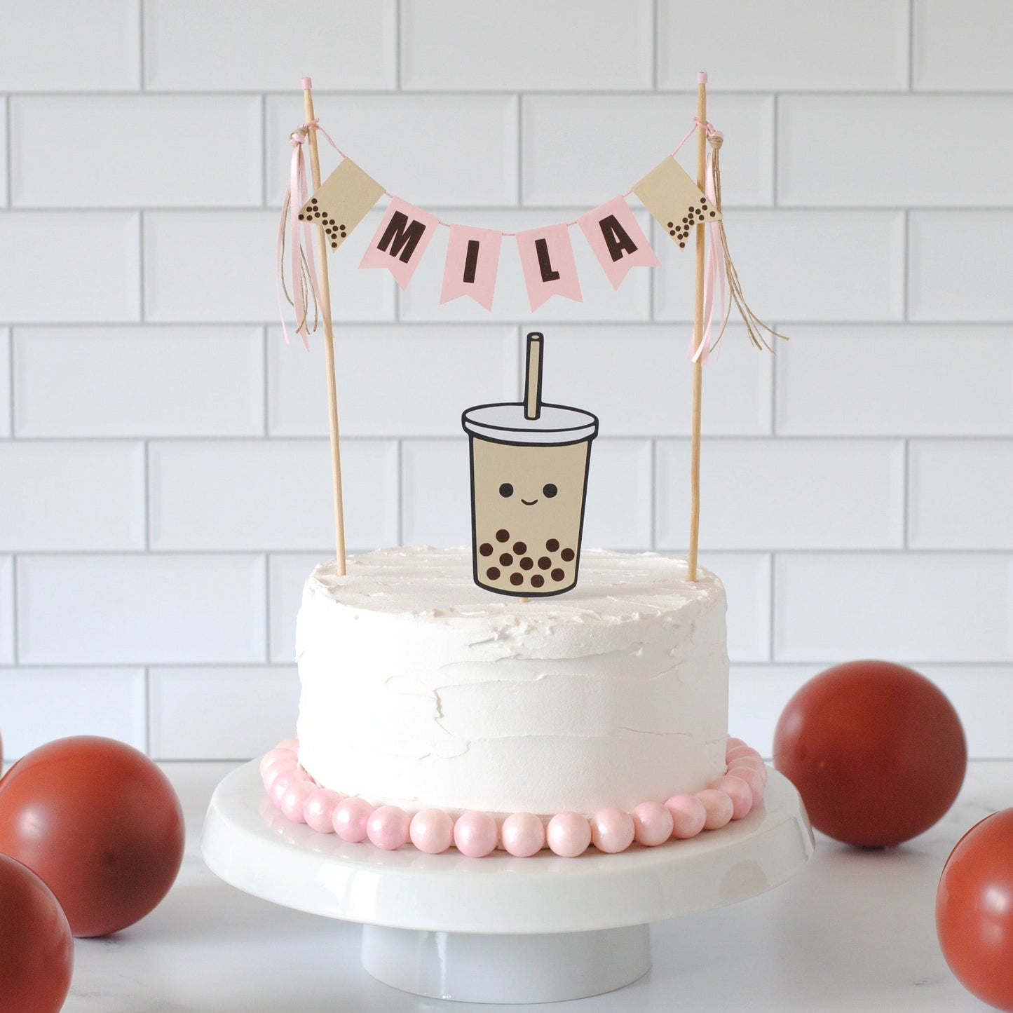 
                  
                    White cake with Boba theme cake topper with name MILA.  Under the banner is a beige Boba tea with a straw. For a Boba theme birthday party
                  
                