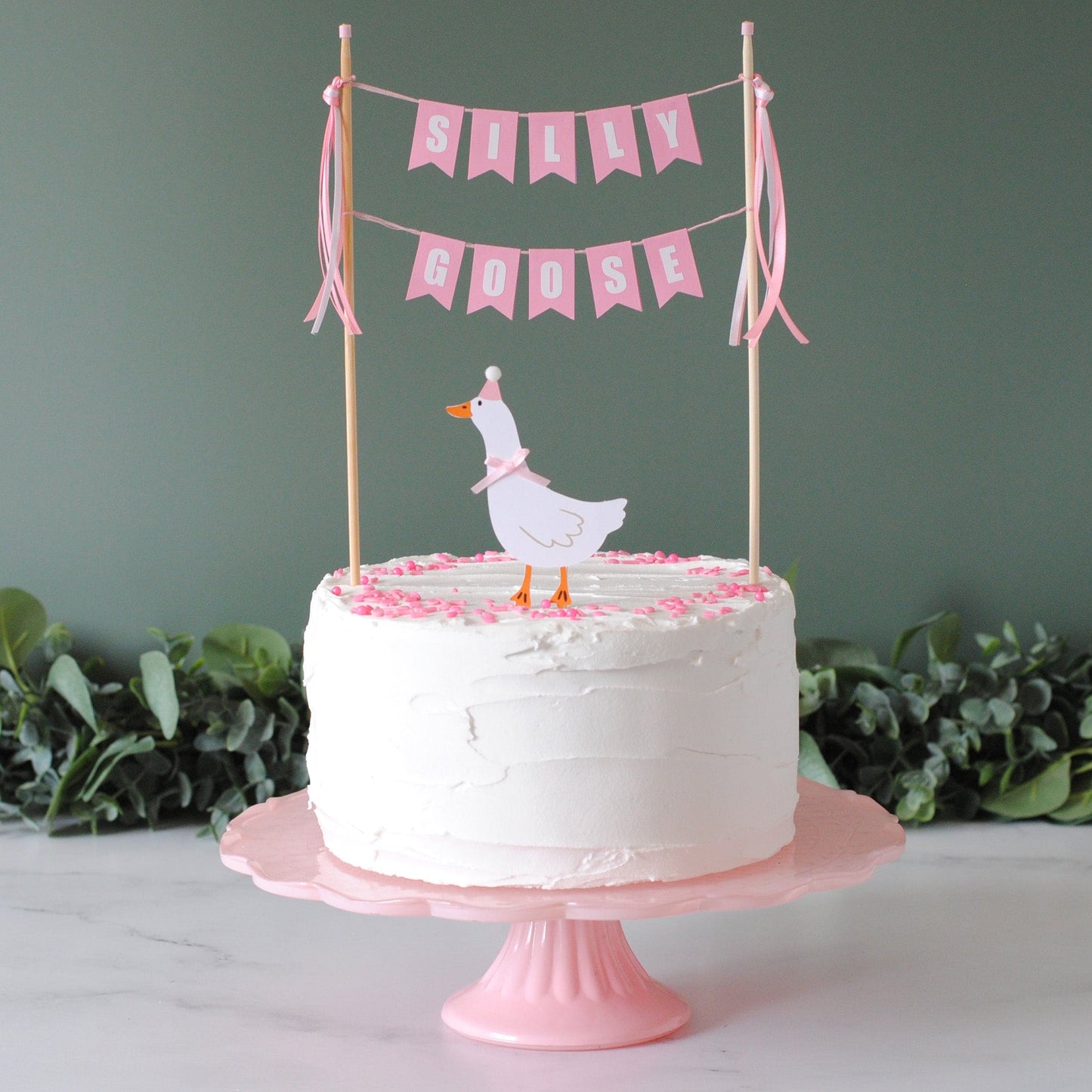 
                  
                    White cake with pink decorations and a goose topper in a party hat with cake banner with 'SILLY GOOSE' on a pink cake stand | cake toppers by Avalon Sunshine
                  
                