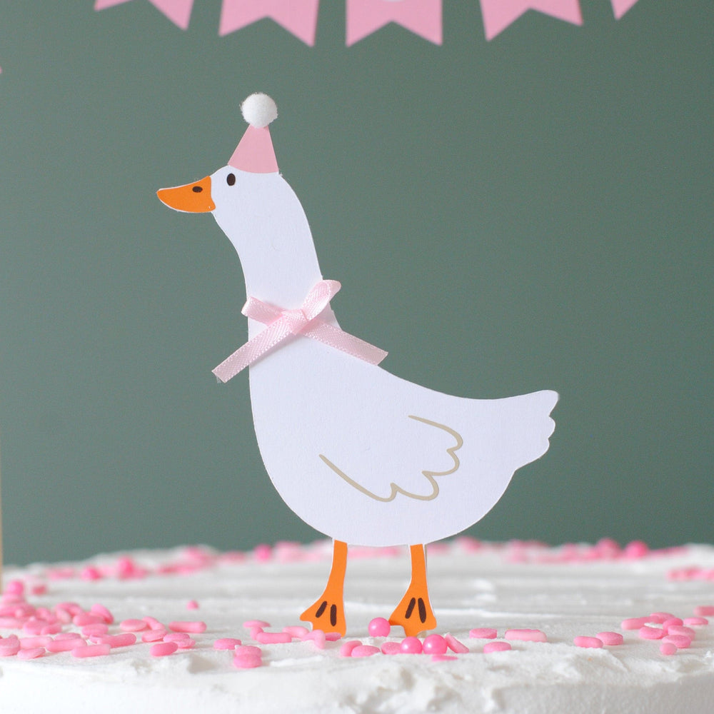 
                  
                    Decorative paper goose with a pink party hat and bow on a cake with pink sprinkles against a green background.
                  
                