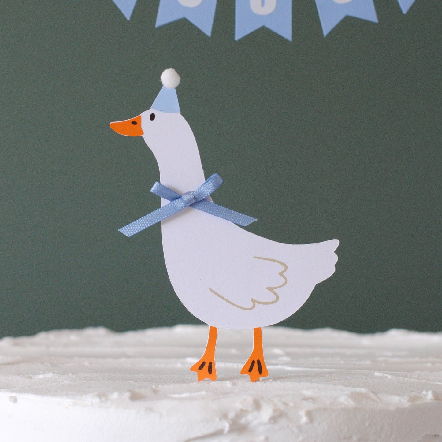 
                  
                    Decorative paper goose with a blue bow and party hat on a cake against a dark green background.
                  
                