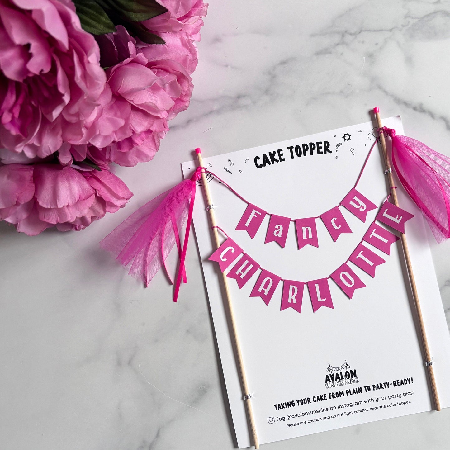 
                  
                    Cake topper with pink tassels and personalized name on a marble surface with pink flowers.
                  
                