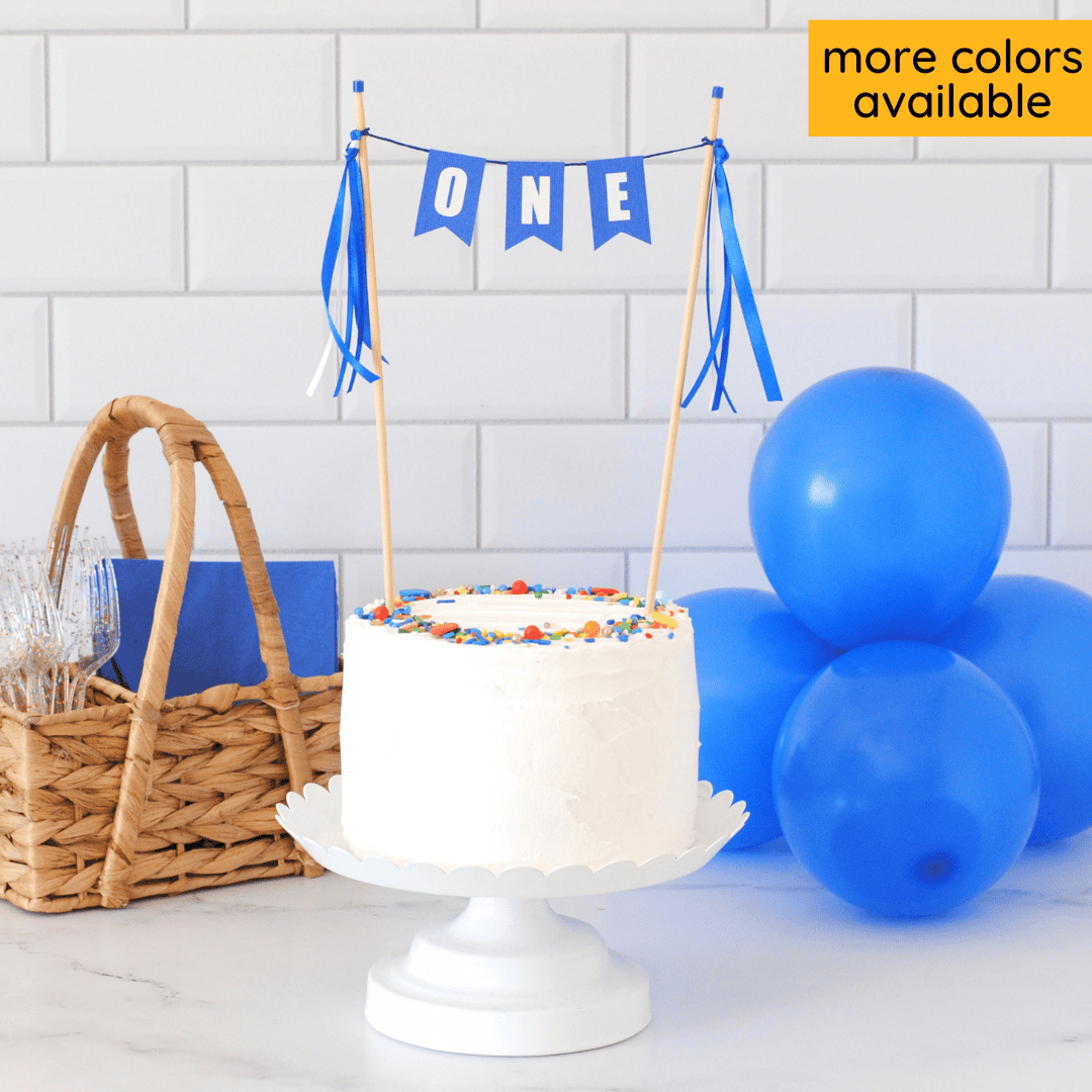ONE birthday cake topper in royal blue with white letters and blue and white ribbon tassels on a small white cake.  Balloons and basket with napkins and forks are in the background