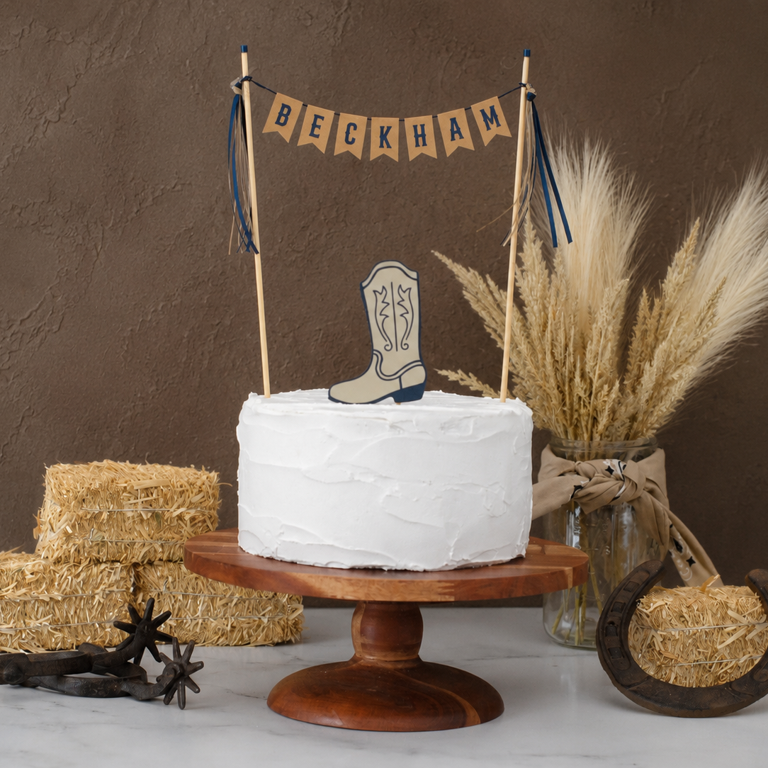 
                  
                    A white frosted round cake on a wooden pedestal stand, decorated with a cowboy boot topper and a banner reading 'BECKHAM'. The setup includes hay bales, a jar of dried florals, a horseshoe, and rustic western decor against a brown textured backdrop.
                  
                