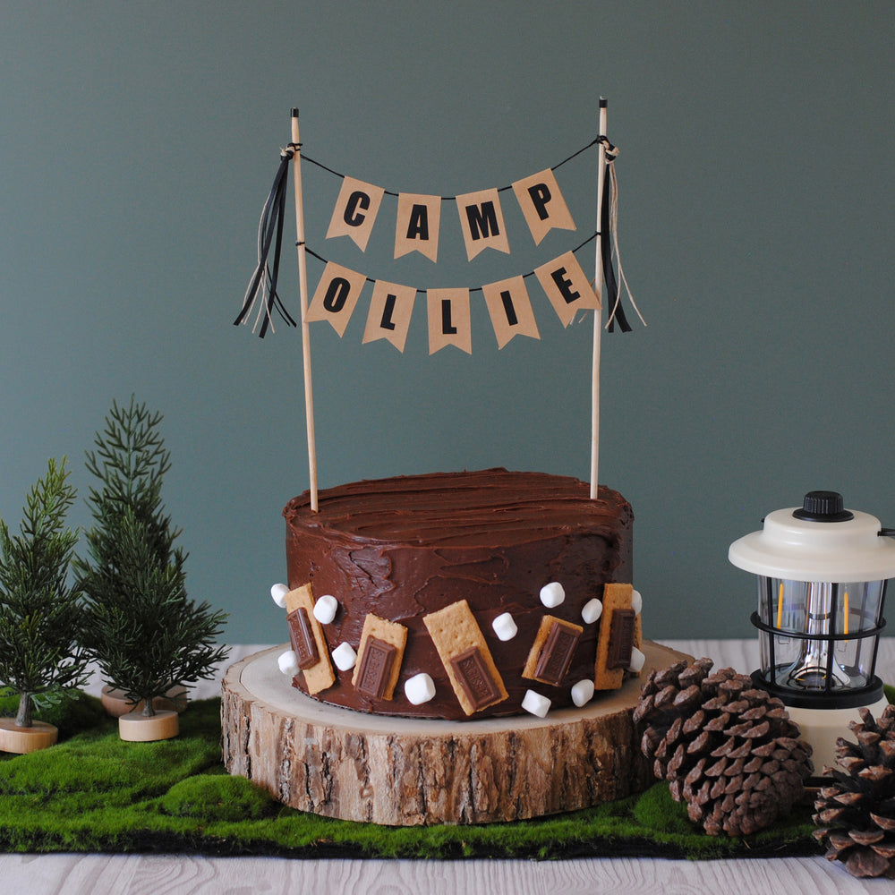 Chocolate cake with s'mores decorations and 'Camp Ollie' Cake Topper against a dark green background | personalized cake toppers by Avalon Sunshine