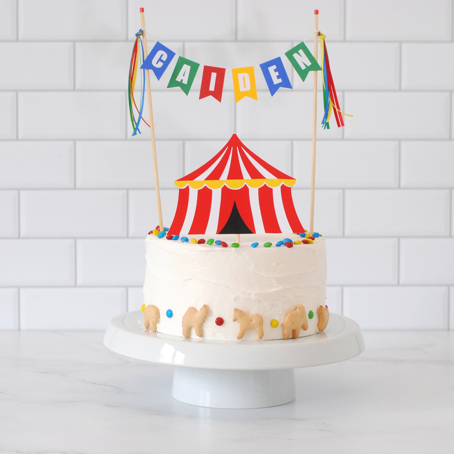 
                  
                    A round white-frosted cake on a white cake stand, decorated with a red-and-white striped circus tent topper, colorful round sprinkles, small animal cookies, and a multicolor banner reading "CAIDEN" above it, set against a white subway-tile background.
                  
                