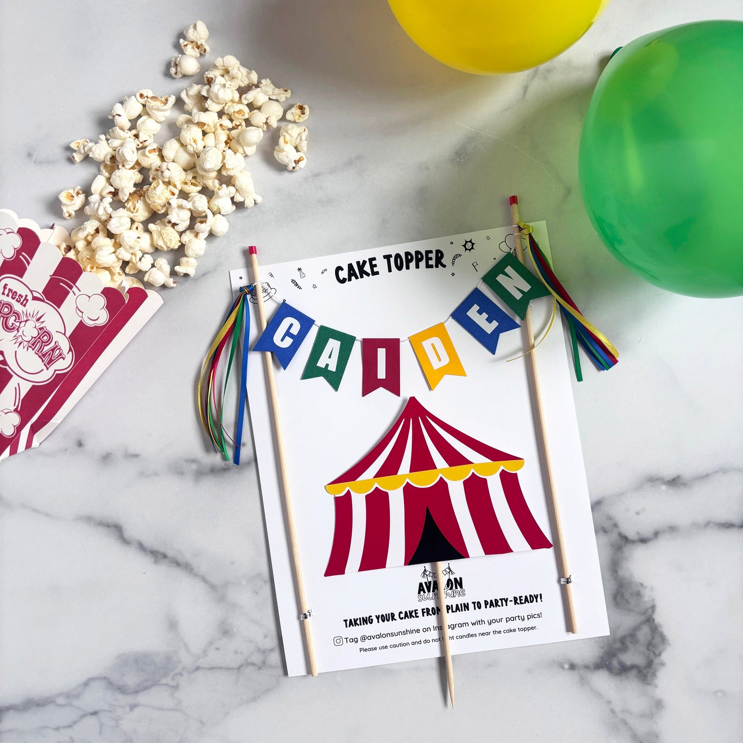
                  
                    A flat-lay of a circus theme cake topper set with name banner and big top on a white marble background, surrounded by party props including popcorn and balloons
                  
                