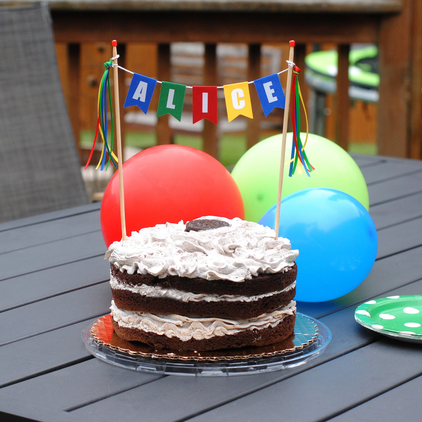 
                  
                    Chocolate cake with white frosting on a table outdoors, decorated with colorful balloons and a 'ALICE' personalized cake topper in bright colors | personalized cake toppers by Avalon Sunshine
                  
                