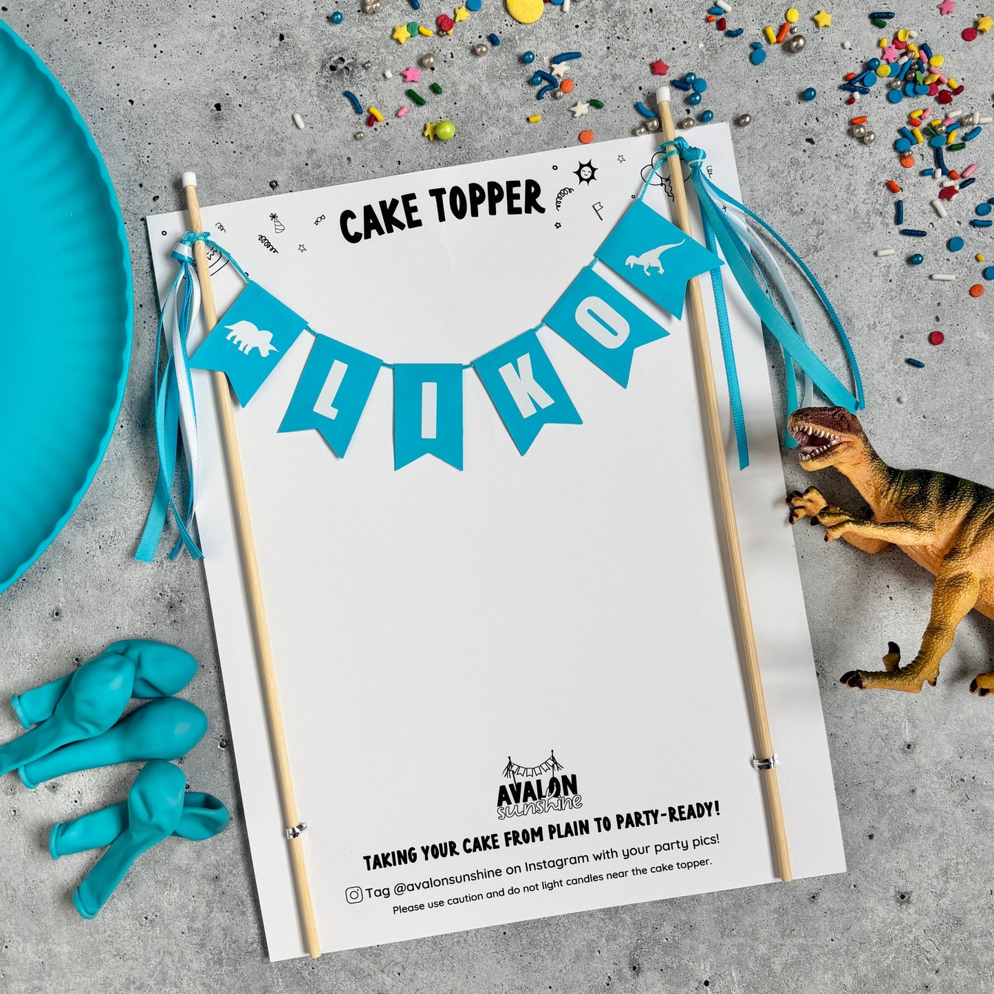 
                  
                    Dinosaur themed cake topper with turquoise flags and wooden sticks personalized with a name.  Shown with sprinkles, balloons and a dinosaur toy.
                  
                