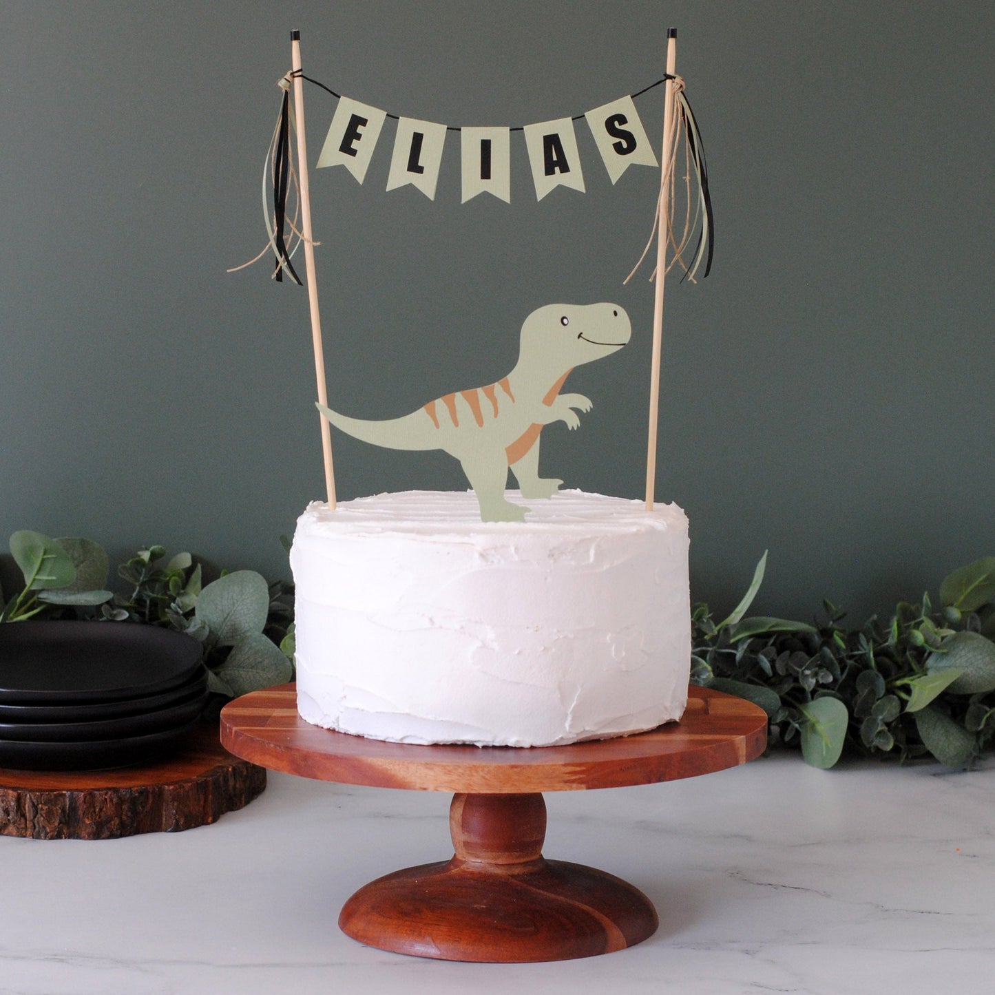 
                  
                    A round white-frosted cake on a wooden pedestal stand, topped with a paper dinosaur topper and a sage green banner reading ELIAS, set against a dark wall with greenery and black plates.
                  
                