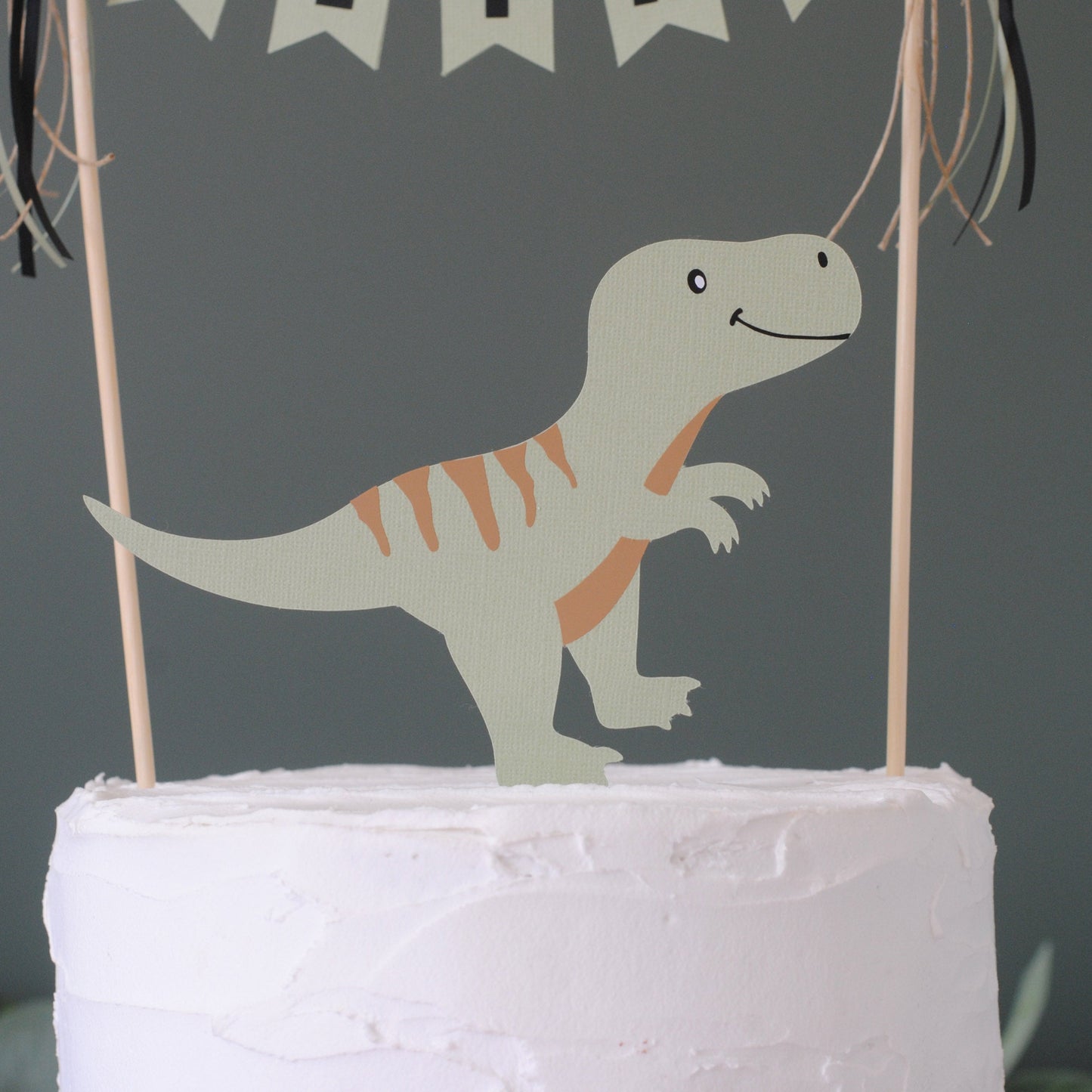 
                  
                    A sage green paper dinosaur cake topper with beige stripes stands on top of a white frosted cake against a dark background.
                  
                