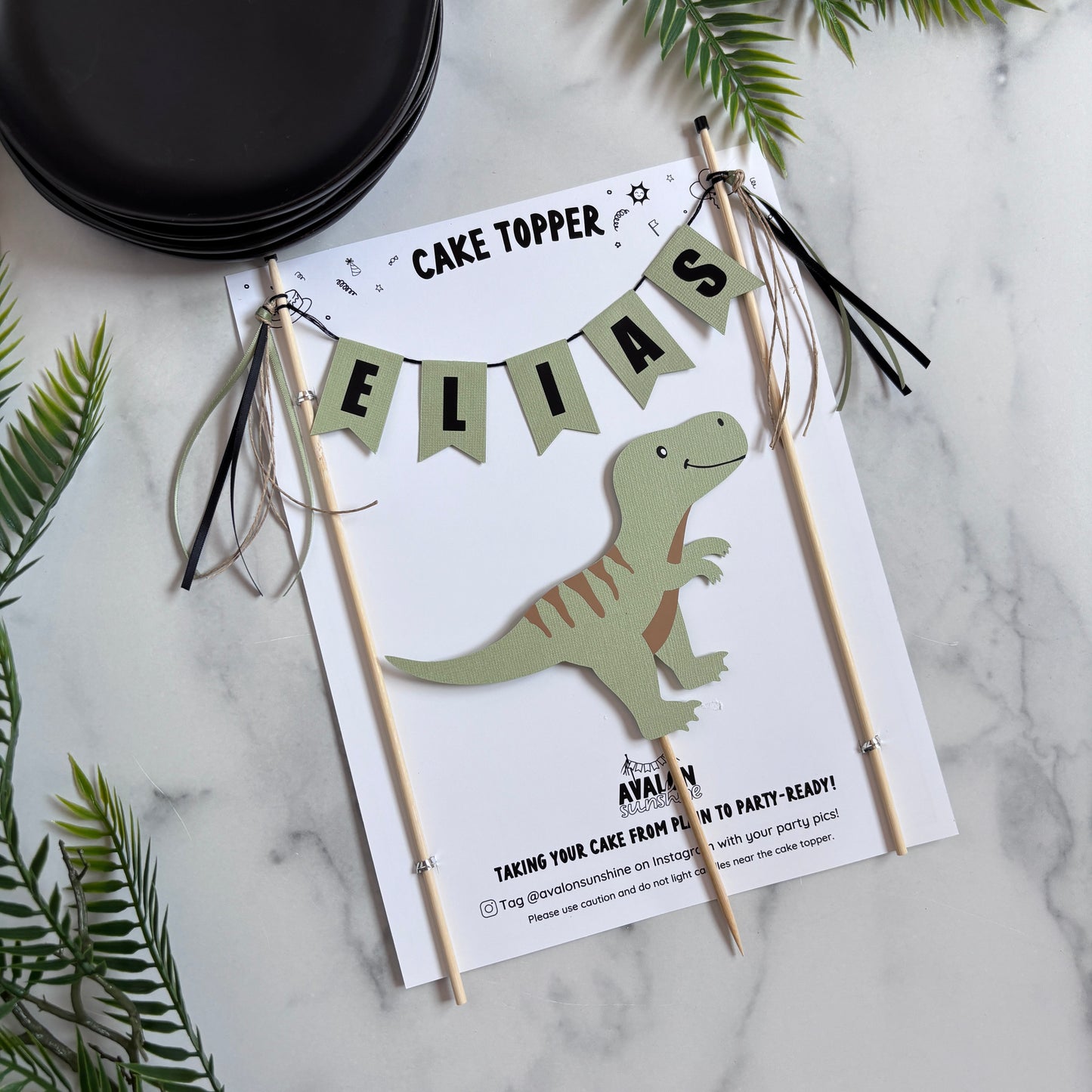 
                  
                    A personalized dinosaur-themed cake topper displayed on a marble surface with green foliage and black plates. The topper features a white card with a sage green dinosaur topper, a sage green banner spelling 'ELIAS', and wooden sticks with ribbon tassels.
                  
                
