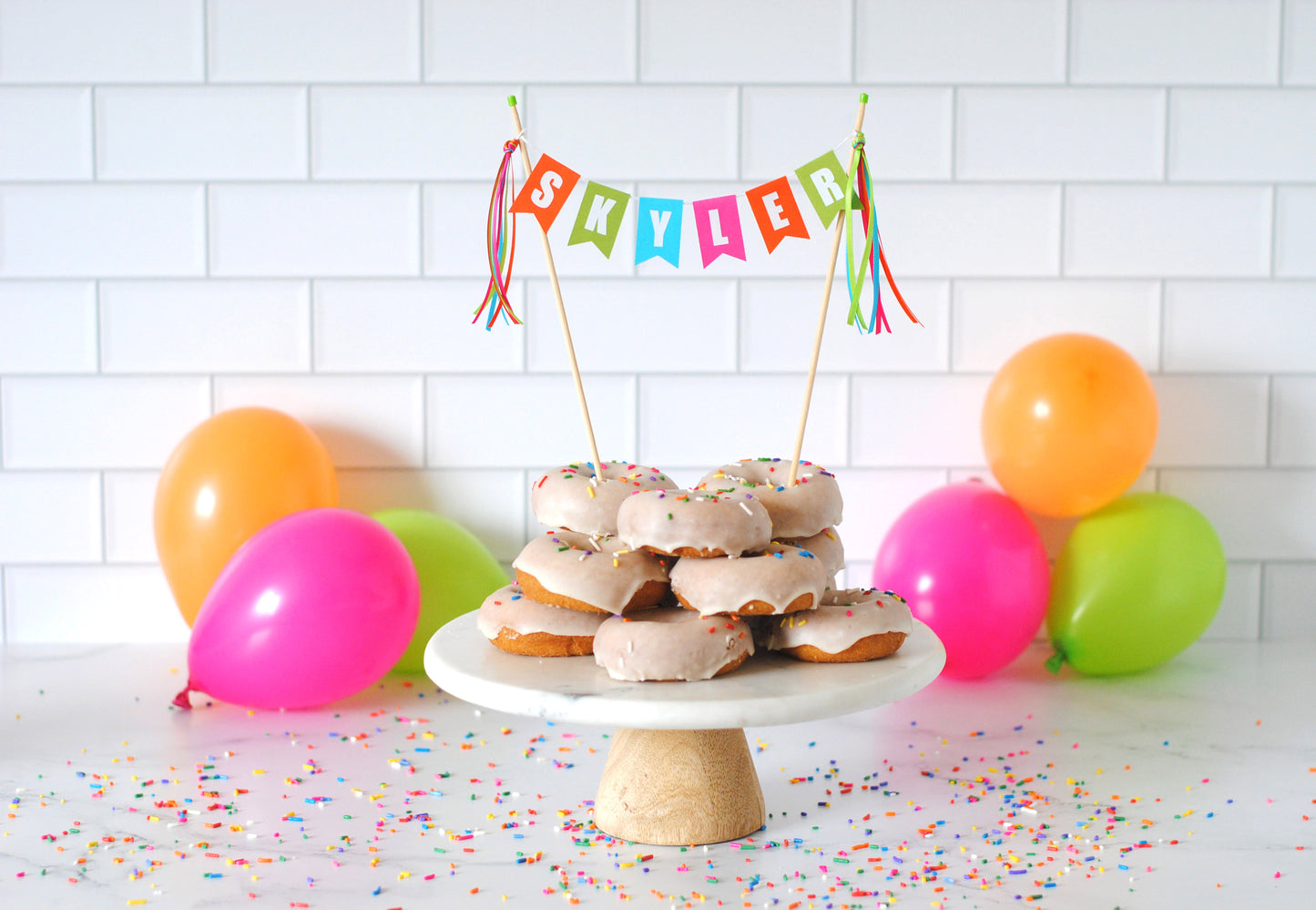 Dozen donuts stacked on a cake stand with name cake topper and balloons in the background | cake topper by Avalon Sunshine