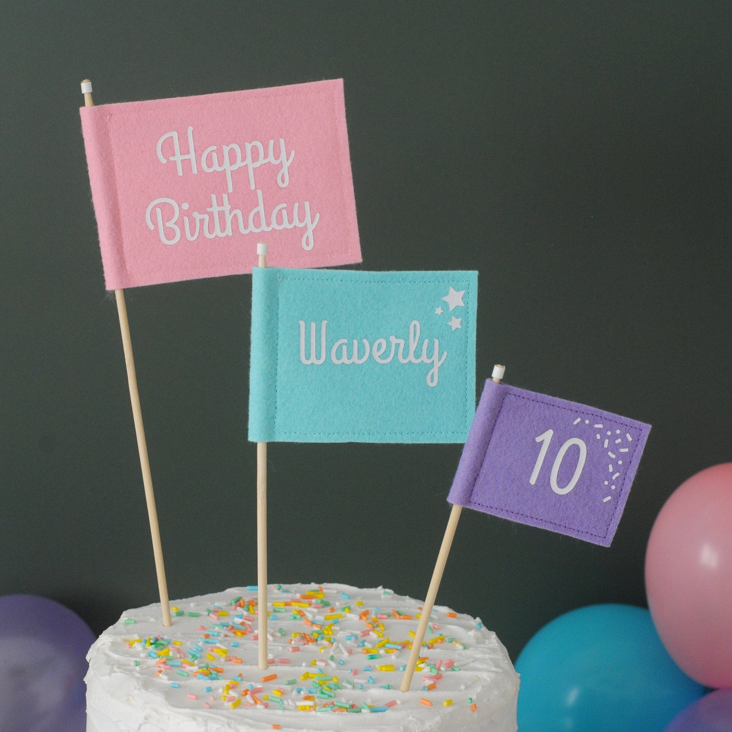 Felt flag cake topper set with pastel colors: large pink HAPPY BIRTHDAY flag, medium aqua blue NAME flag, and small purple AGE flag 