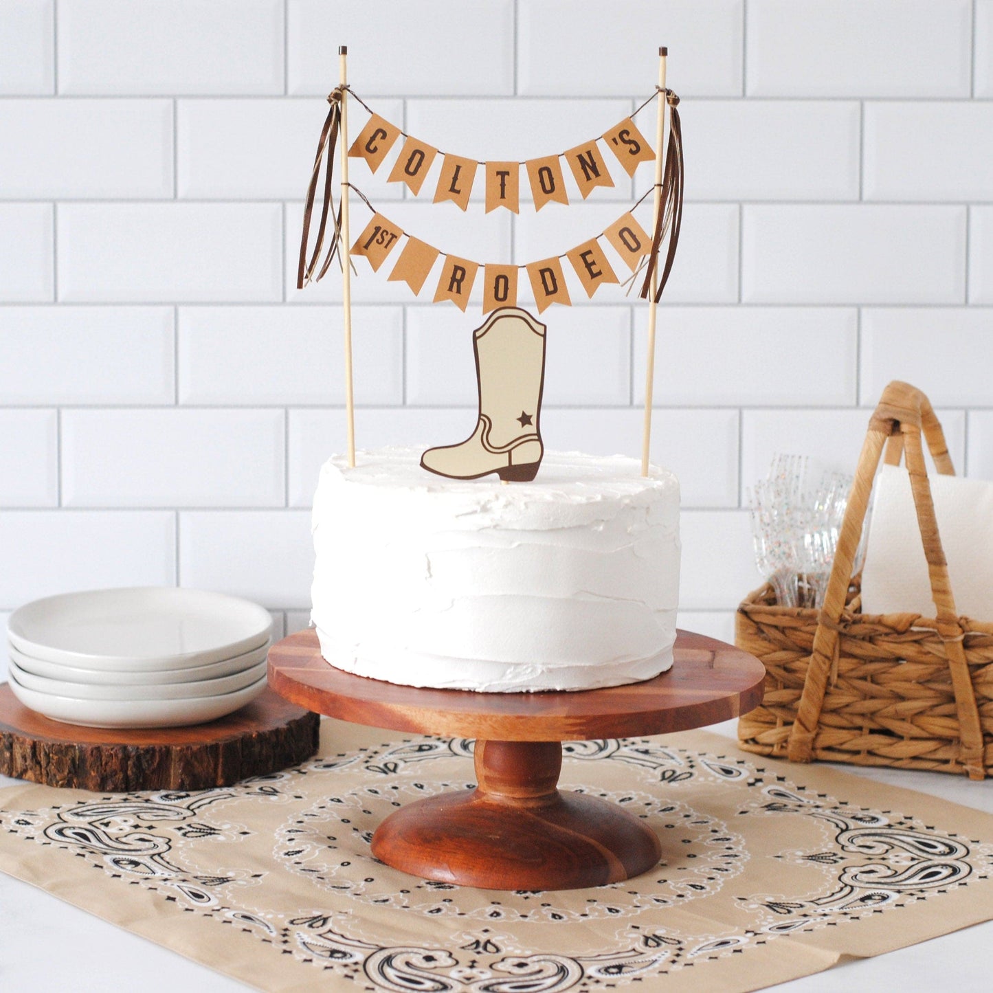
                  
                    White cake on a wooden stand with a 'COLTON'S 1ST RODEO' banner and cowboy boot topper, set against a white tiled wall.
                  
                