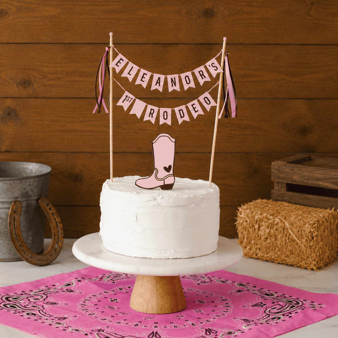 
                  
                    White cake with a pink boot topper and 'Eleanor's 1st Rodeo banner on a cake stand, with a pink bandana and rustic items in the background.
                  
                
