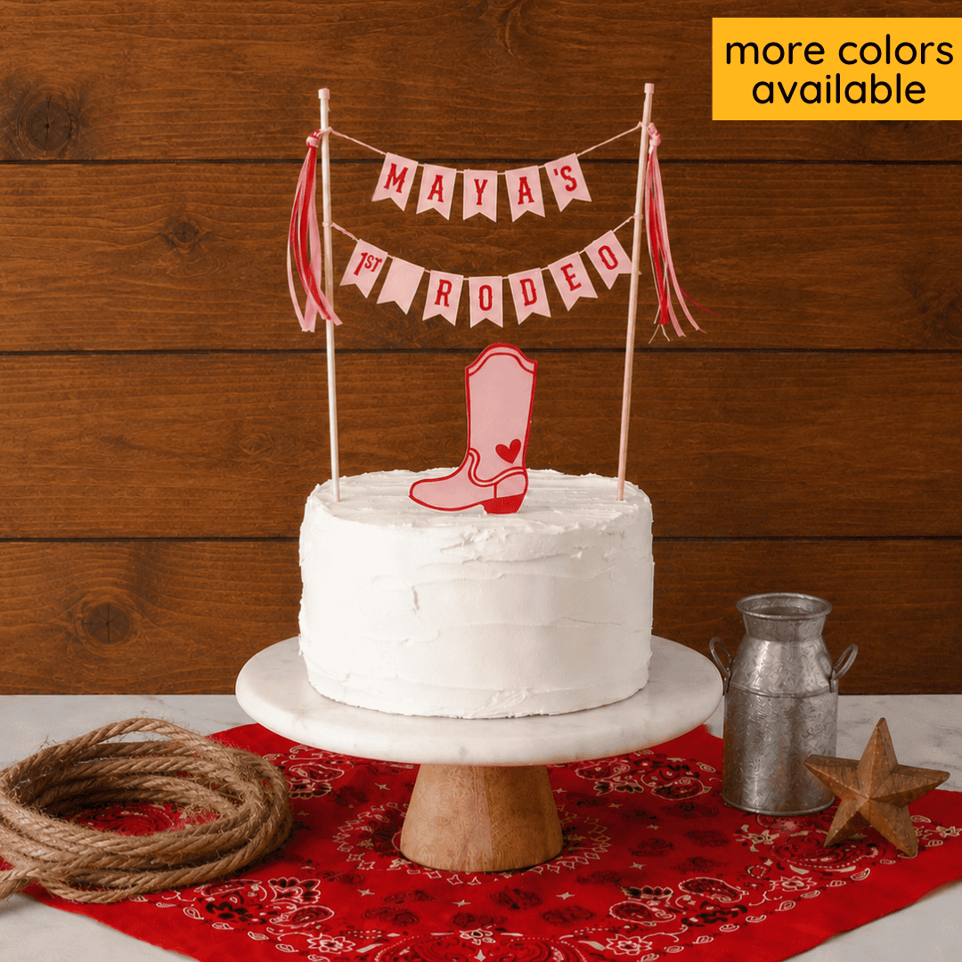 White cake with cowboy boot topper and 'MAYA'S 1st RODEO' banner on a wooden background