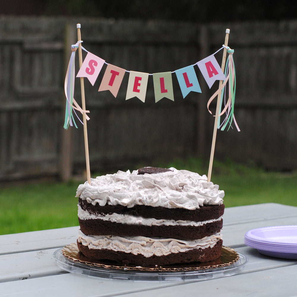 
                  
                    Chocolate cake with white frosting on a picnic table, decorated with a 'STELLA' banner in pastel rainbow colors | personalized cake topper by Avalon Sunshine
                  
                