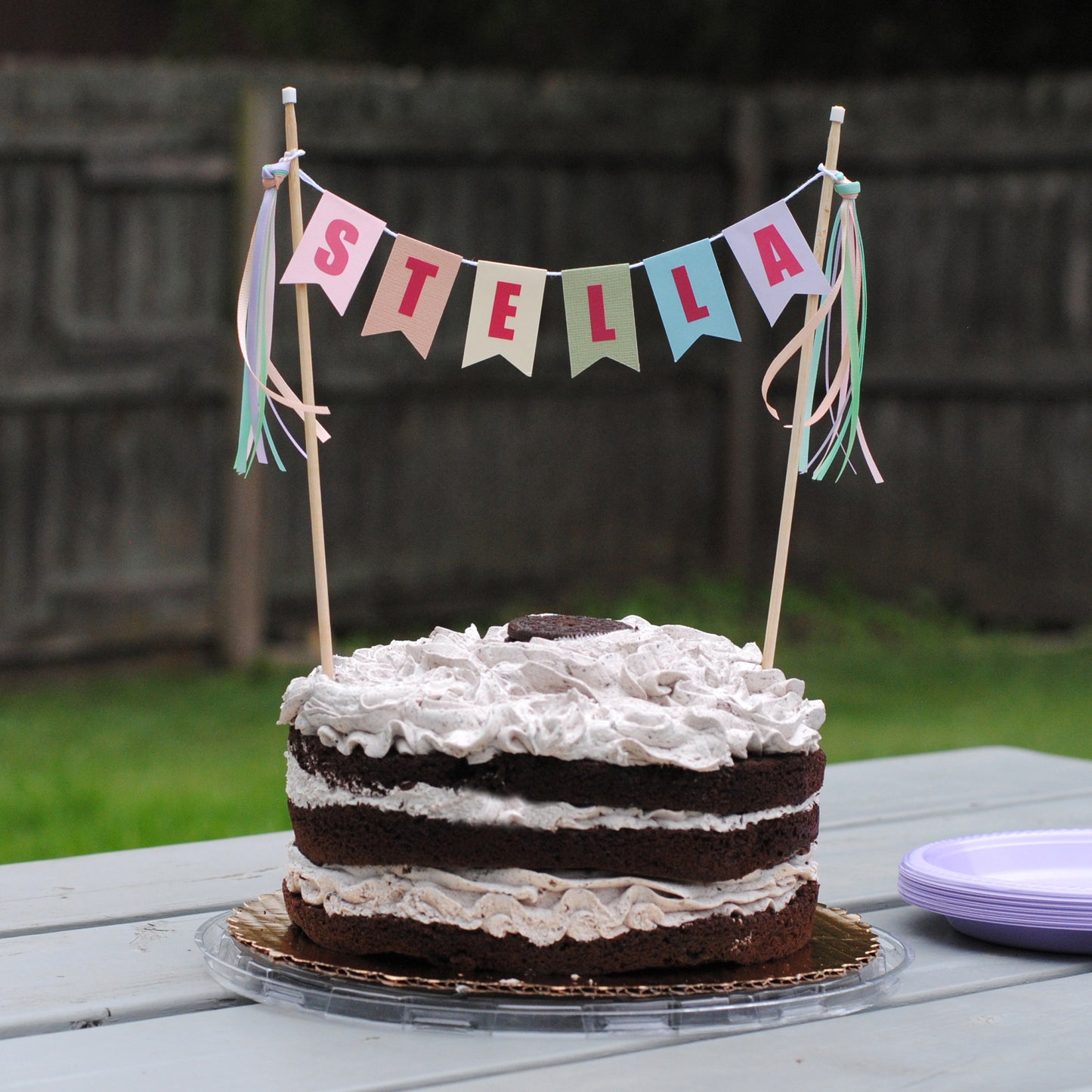 
                  
                    Chocolate cake with white frosting on a picnic table, decorated with a 'STELLA' banner in pastel rainbow colors | personalized cake topper by Avalon Sunshine
                  
                