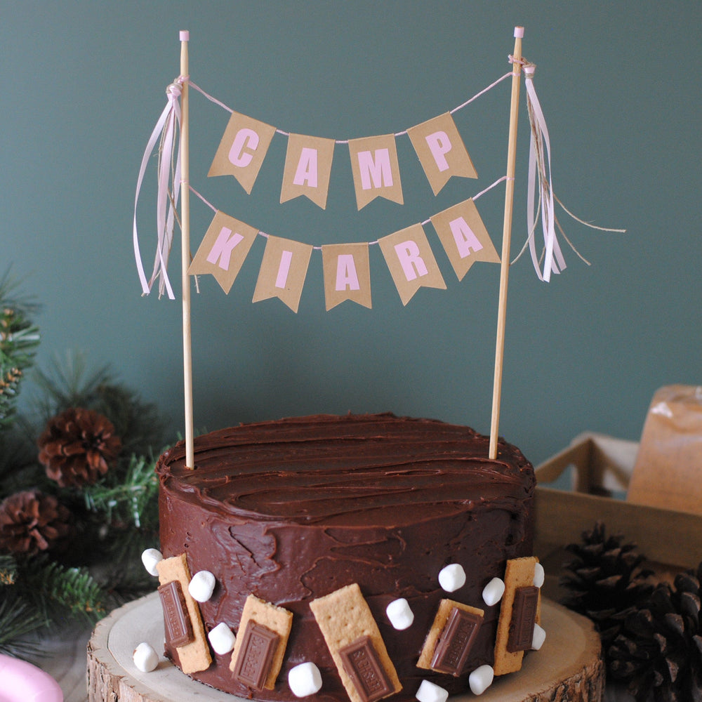 Chocolate cake with s'mores decorations, topped with a 'Camp Kiara' cake topper with pink letters | personalized cake toppers by Avalon Sunshine