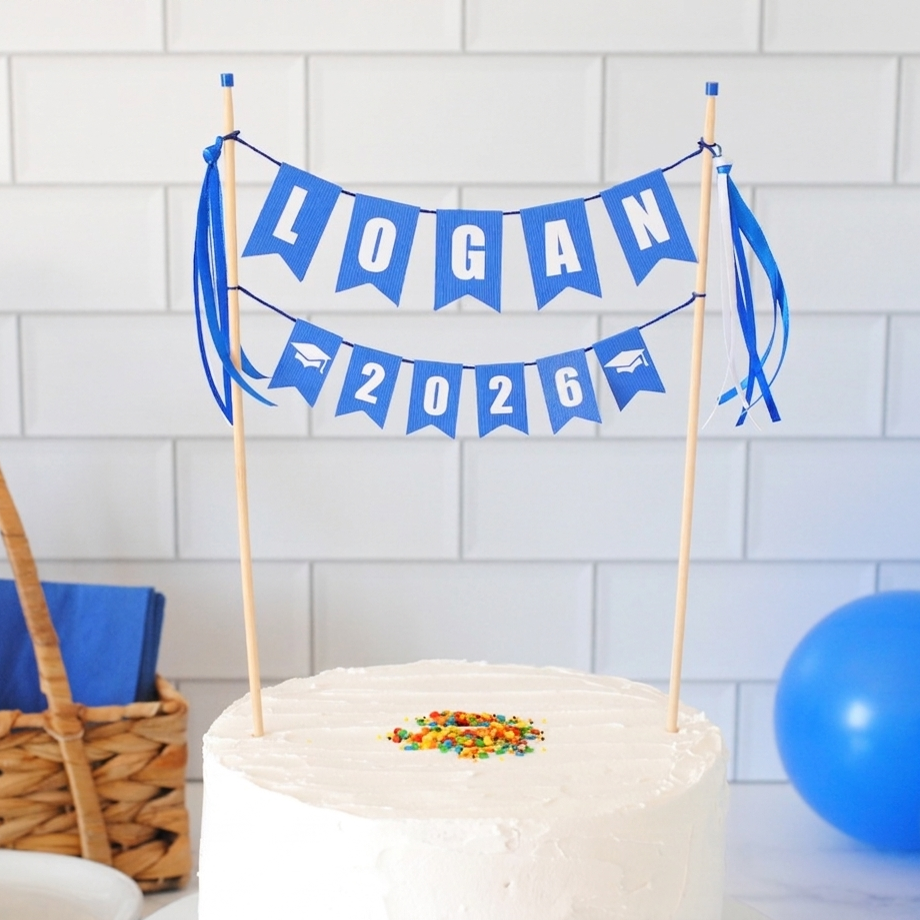 
                  
                    2026 Graduation cake topper: White cake with colorful sprinkles, blue 'LOGAN 2026' banner, and blue balloon against a white tiled wall.
                  
                