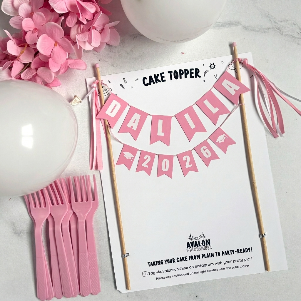
                  
                    Graduation 2026 Cake topper with pink banner personalized with name and grad caps on a white surface with pink flowers and forks.
                  
                
