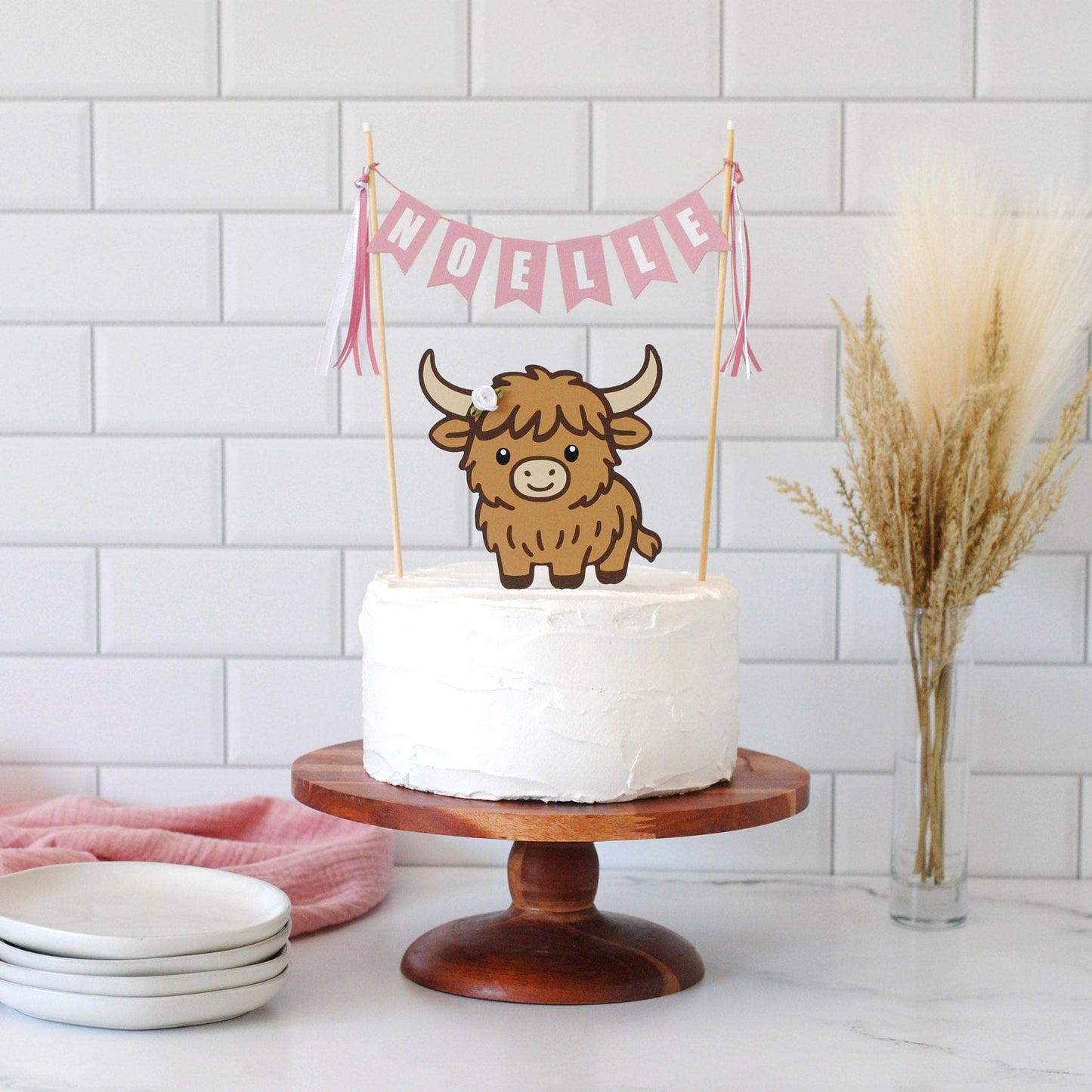 A white frosted round cake on a wooden pedestal stand, decorated with a pink name banner topper and a brown Highland cow topper. The cake is set on a marble countertop with a white subway tile backsplash, a stack of white plates on the left, and a vase of dried pampas grass on the right.