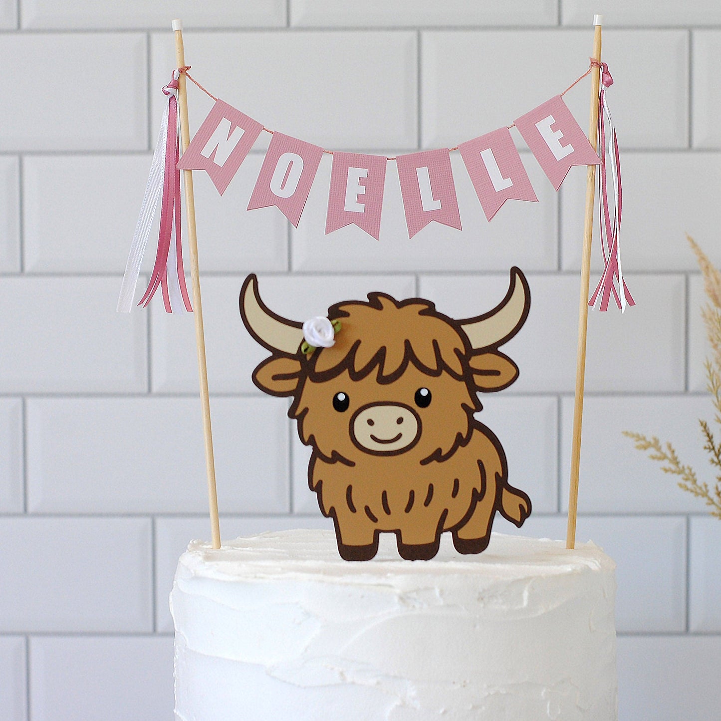 
                  
                    A Highland cow cake topper on a white frosted cake, featuring a pink bunting banner that reads 'NOELLE' with pink and white tassels, set against a white subway tile background.
                  
                
