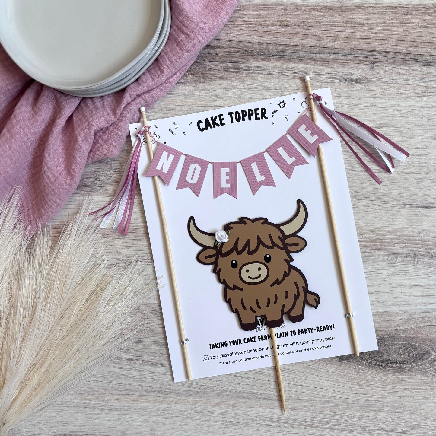 
                  
                    A styled flat-lay of a custom 'Cake Topper' sign featuring a brown Highland cow and a pink bunting banner spelling 'NOELLE' on wooden sticks, set on a light wood surface with a pink cloth and plates in the background.
                  
                