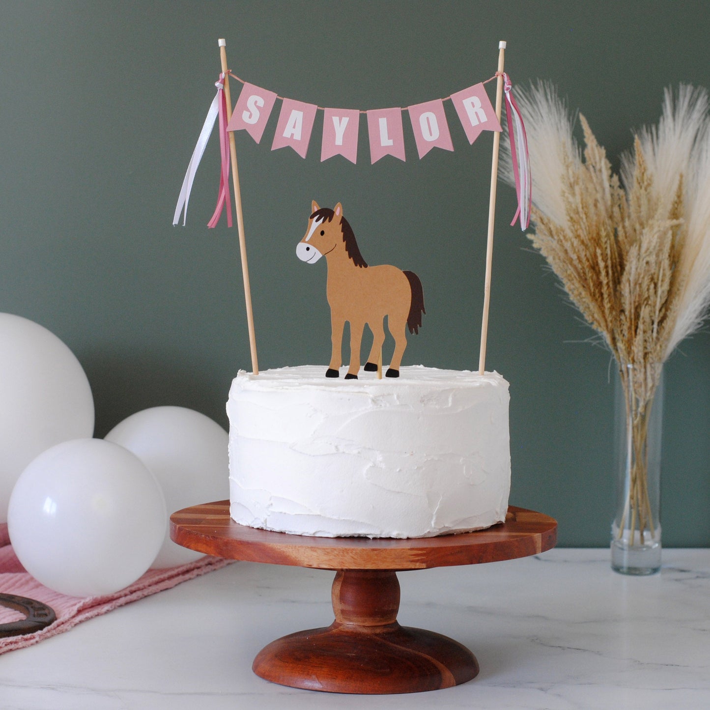 
                  
                    A white frosted round cake on a wooden pedestal stand, decorated with a pink banner reading 'SAYLOR' and a brown horse topper. The scene is styled with white balloons, a pink cloth, and dried pampas grass against a dark green wall.
                  
                