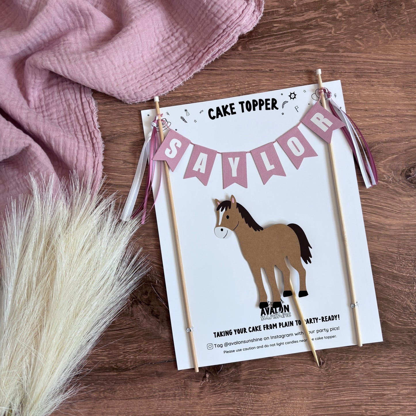 
                  
                    A horse cake topper set with a name banner in dusty pink color and a separate brown horse topper.  displayed in packaging against a wood background
                  
                