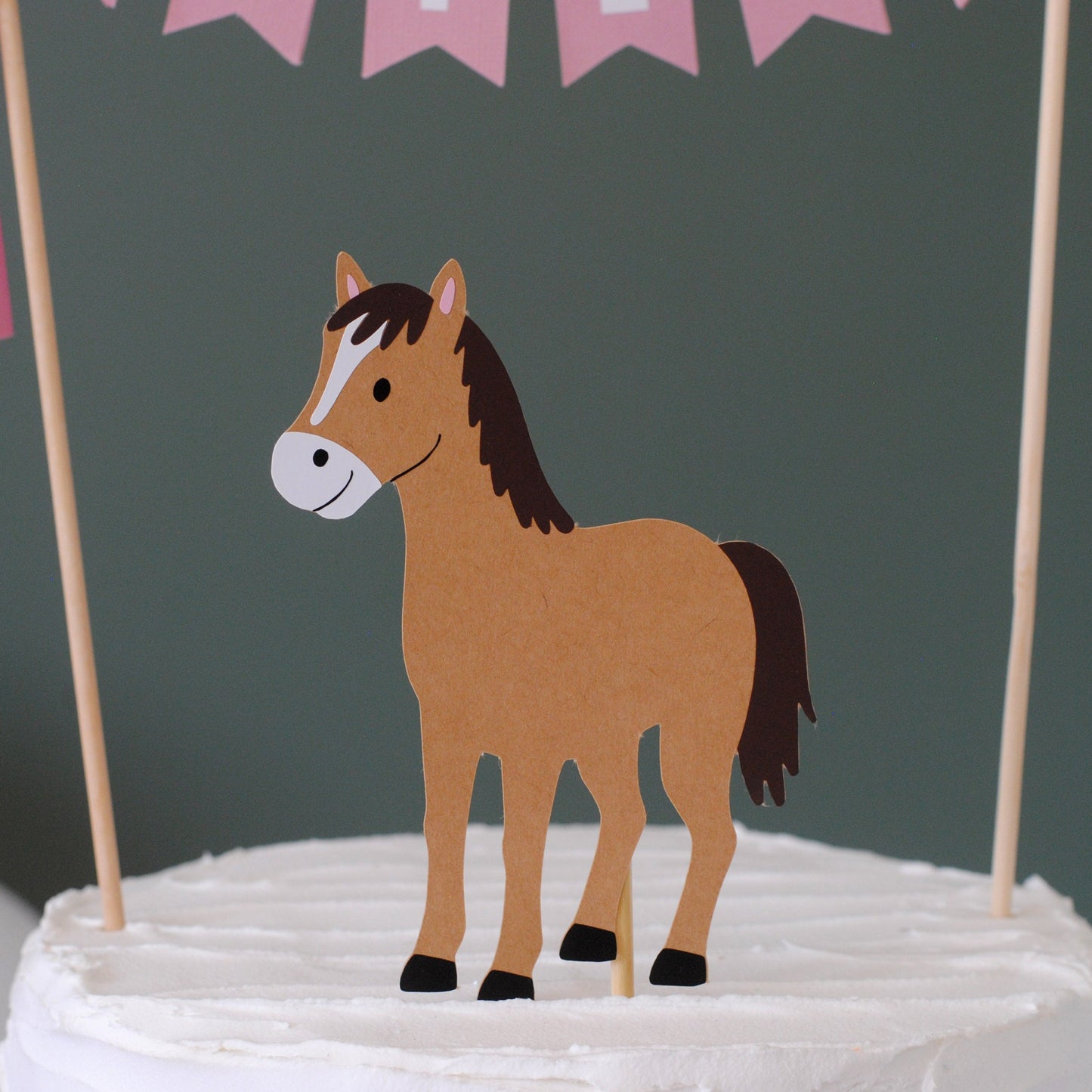 
                  
                    A brown paper cutout horse cake topper with a white snout and black hooves, displayed on a white frosted cake with pink and white streamers against a dark green background.
                  
                