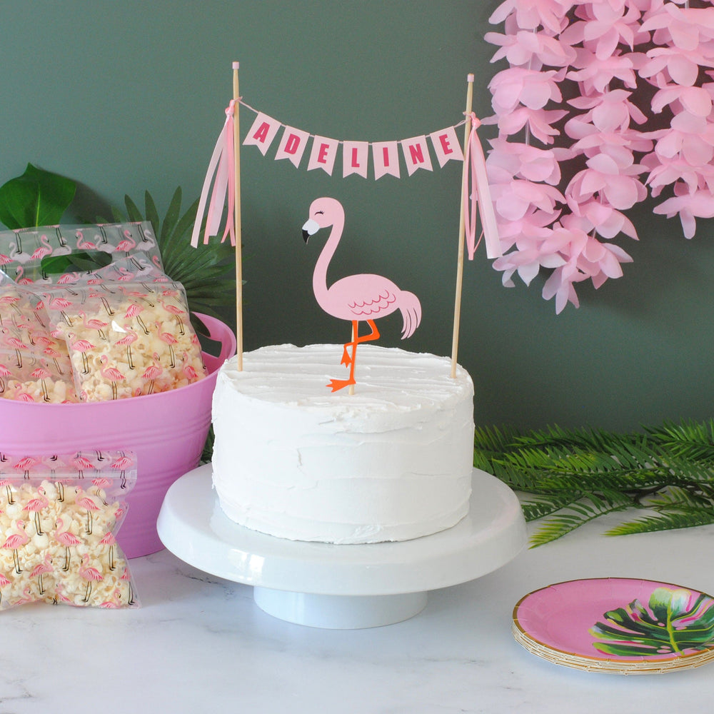 pink flamingo cake topper with a personalized pink name banner on the cake.  Shown on a white cake with pink flower lei in the background and a pink bin with flamingo treat bags | personalized cake toppers by Avalon Sunshine