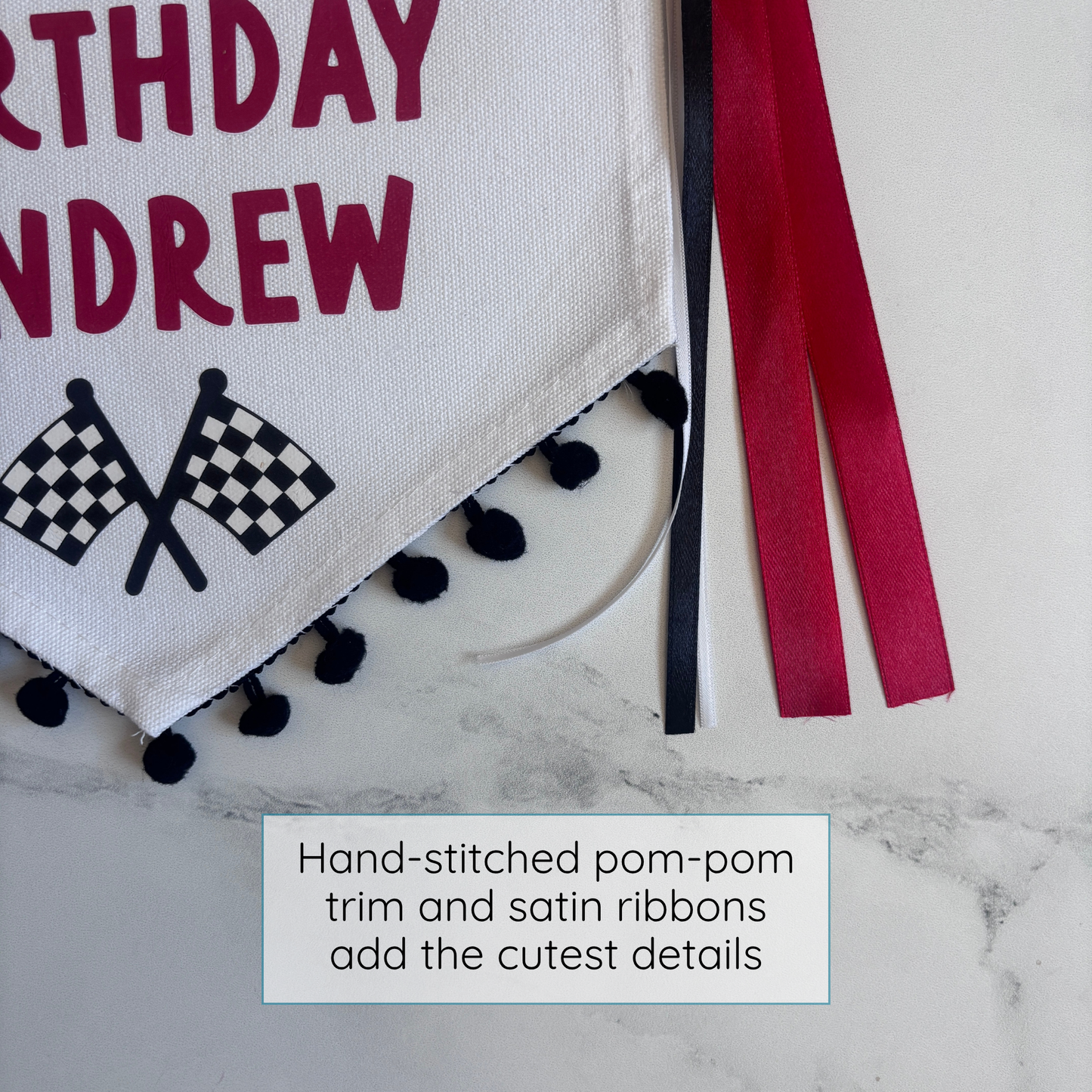 
                  
                    Decorative banner with 'Happy Birthday Andrew' text, checkered flags, pom-pom trim, and satin ribbons on a marble surface.
                  
                